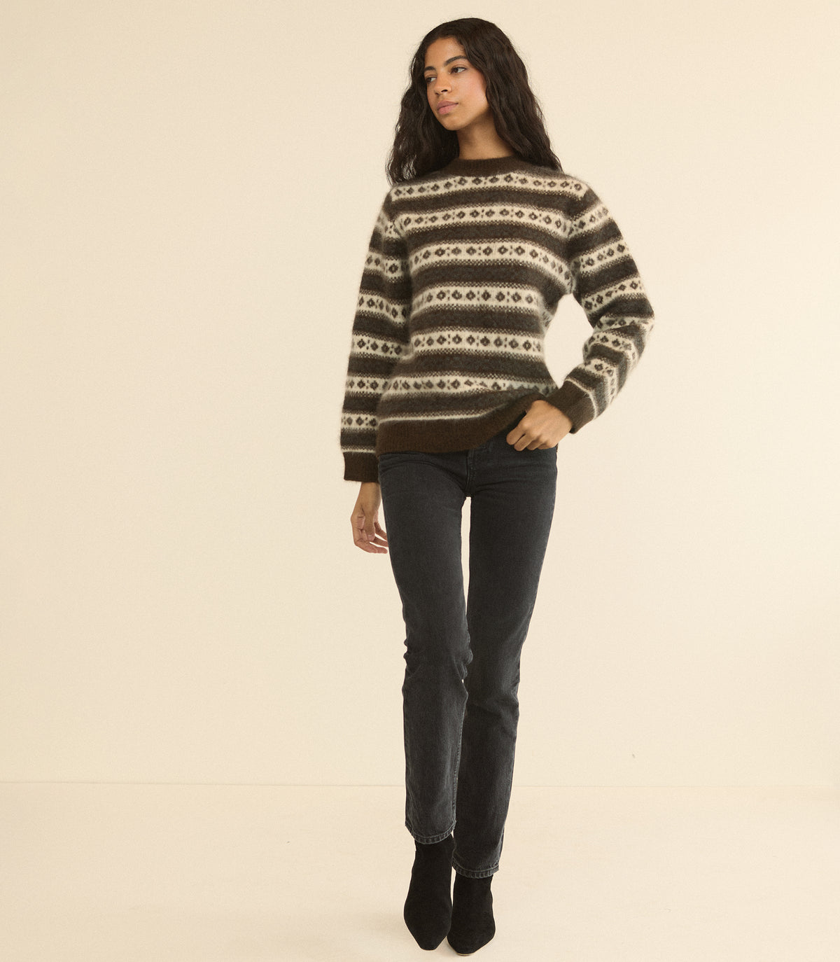 A woman modeling a brown and beige patterned knit sweater with dark gray jeans and black ankle boots, standing against a solid cream background.