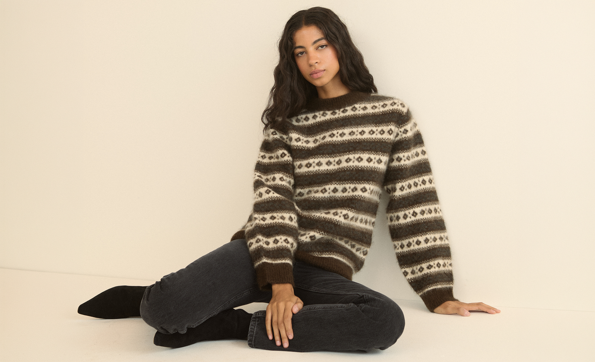 A woman sits on the floor against a cream wall wearing a brown and cream Fair Isle patterned crewneck sweater with dark gray jeans and black ankle boots.
