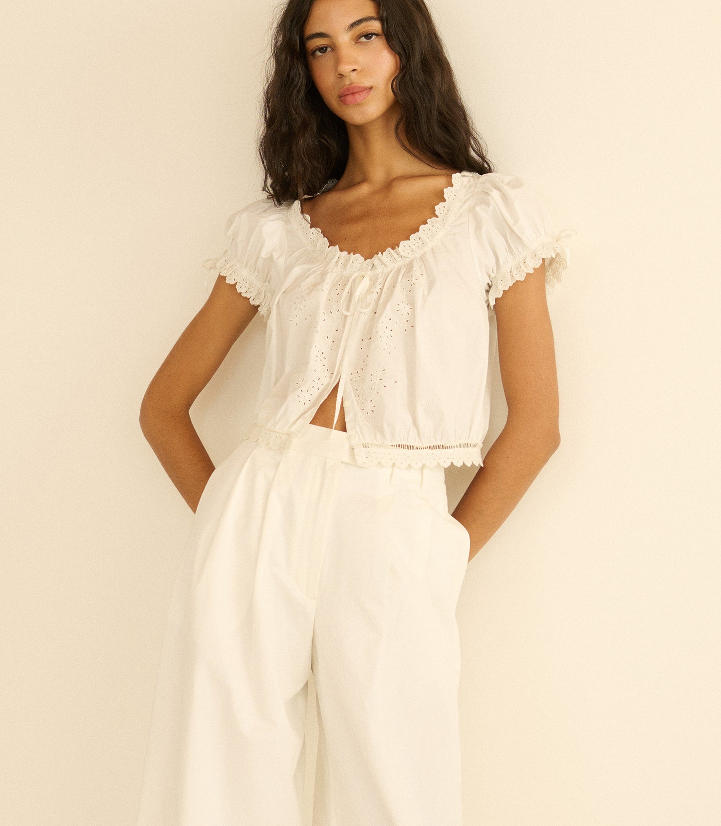 Woman wearing a white outfit with ruffled top and wide-leg pants against a beige background