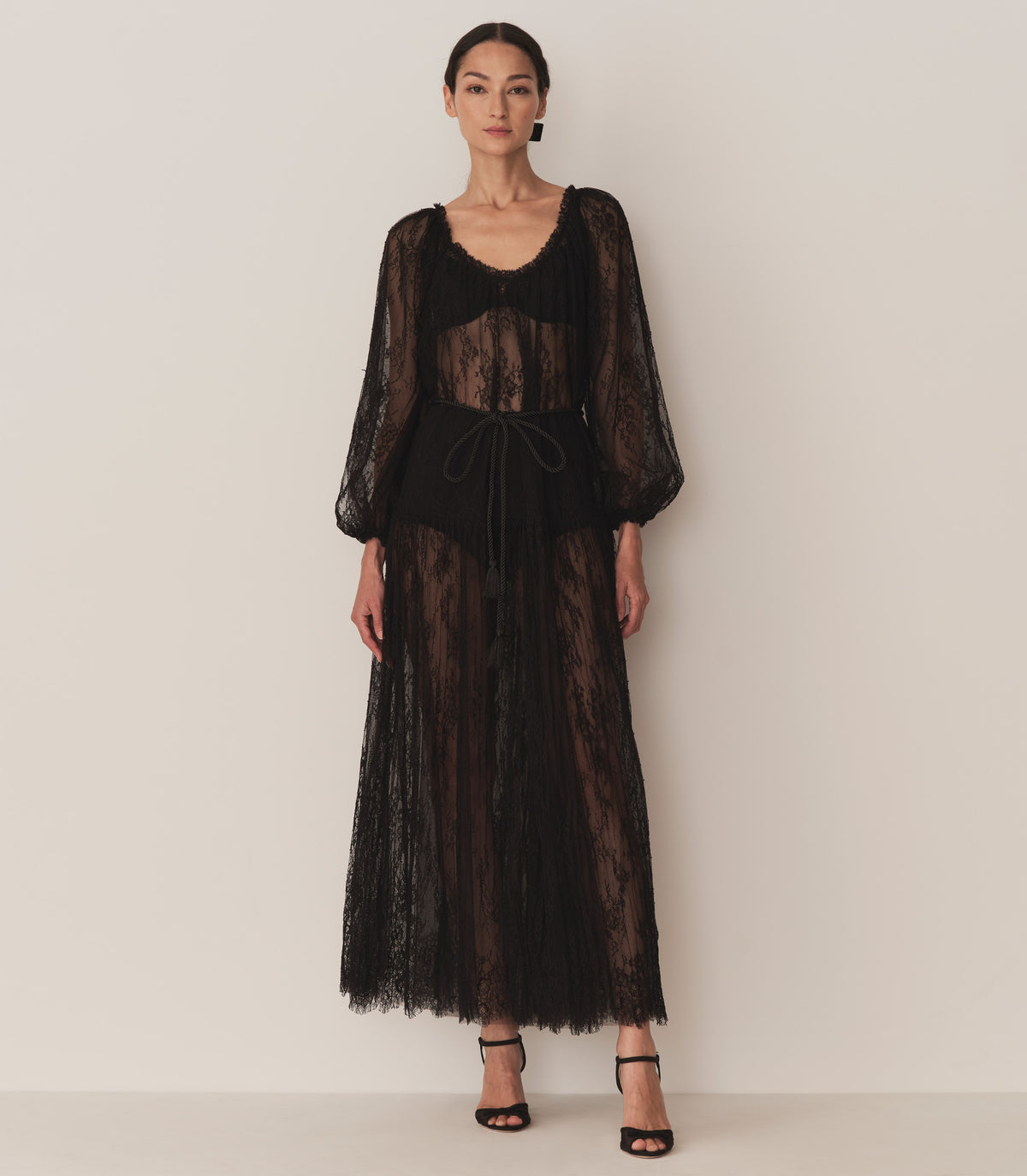 A woman wearing the Nevaeh Dress, a long black sheer lace dress with long sleeves and a tie at the waist.