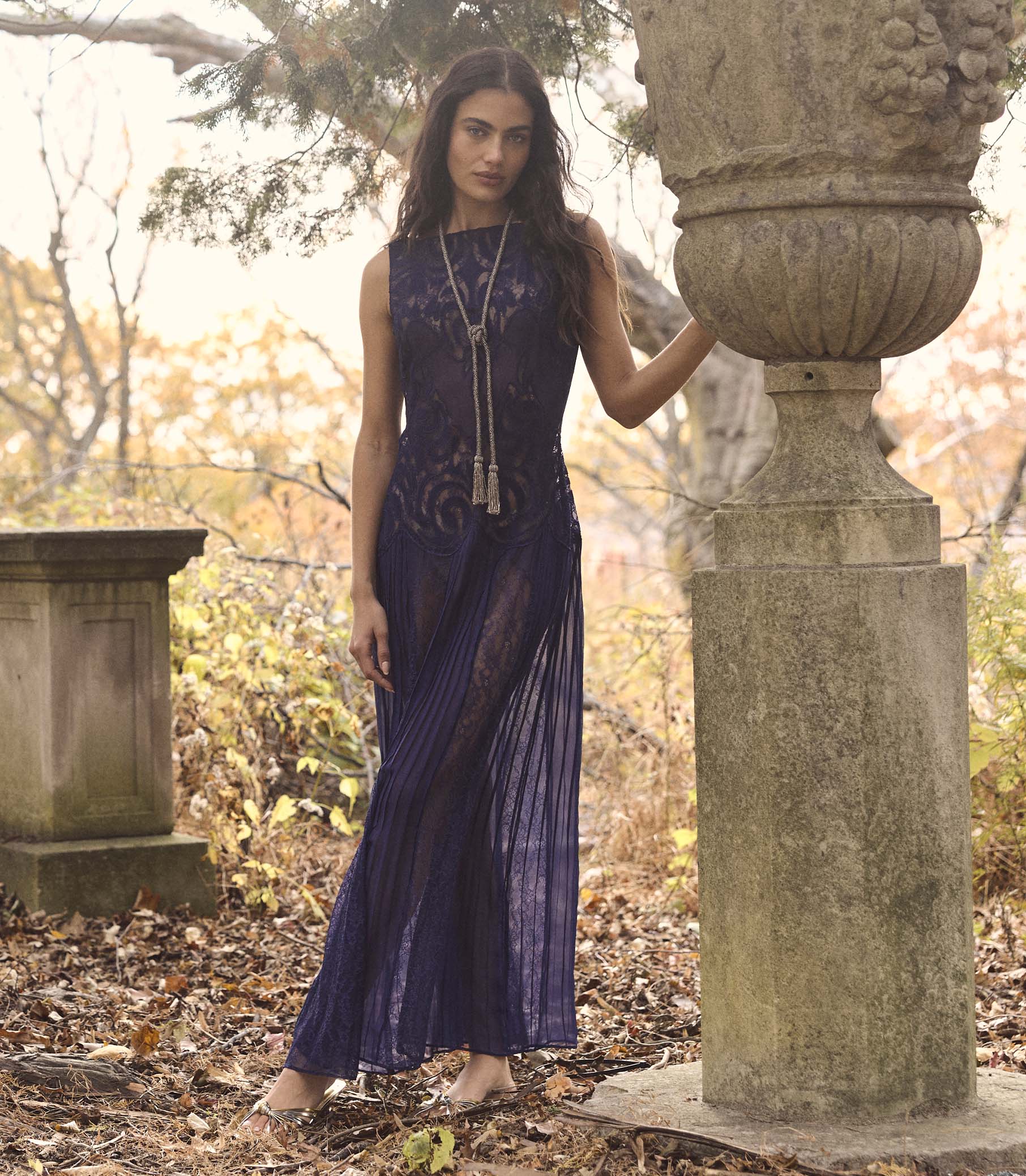 Woman in a purple dress standing in an outdoor setting with stone structures and foliage.