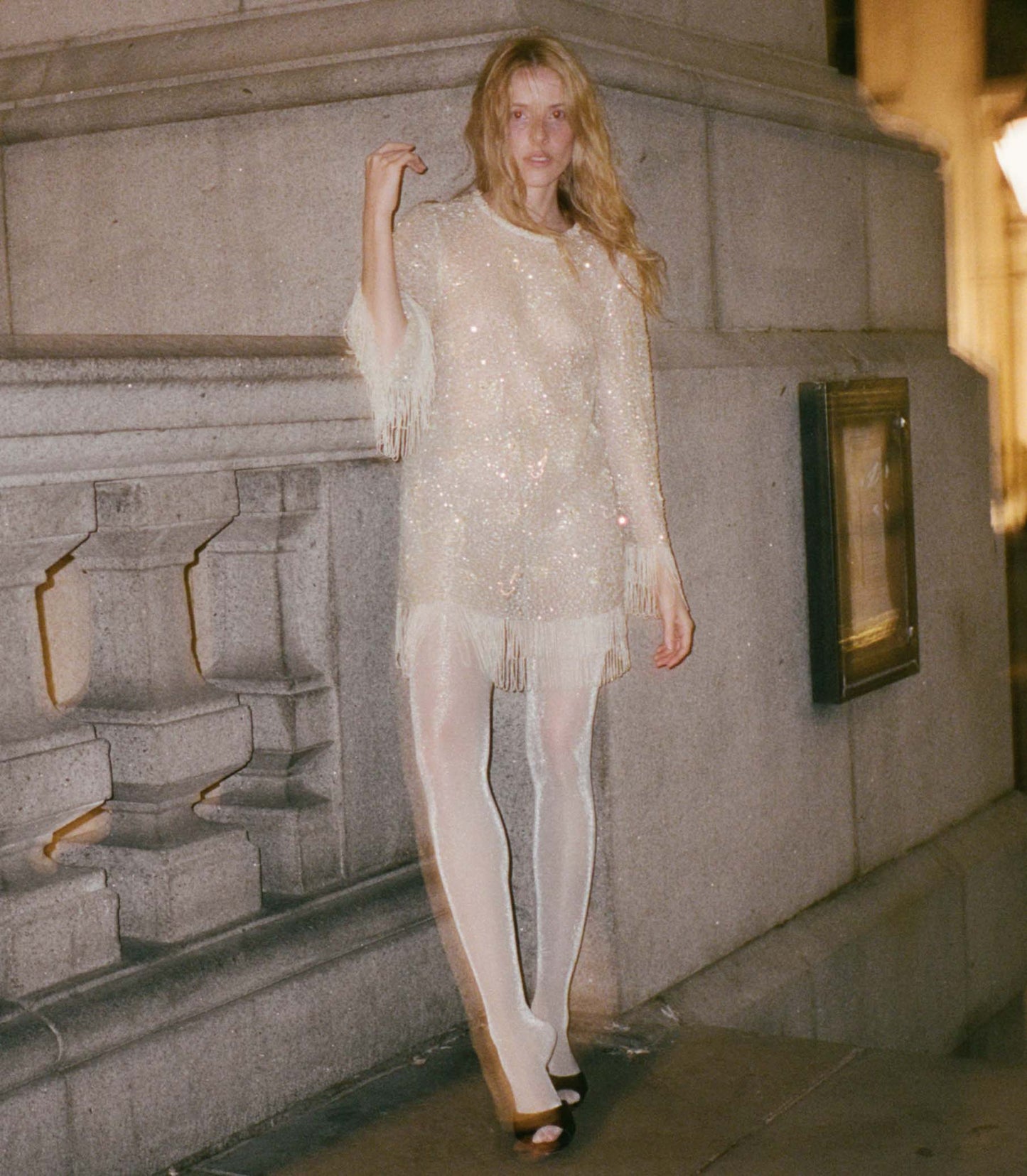 Woman in a light shimmering fringed mini dress standing against a stone wall.