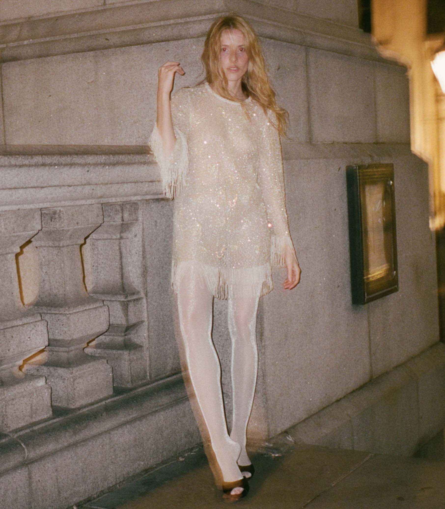 Woman in a light shimmering fringed mini dress standing against a stone wall. 1