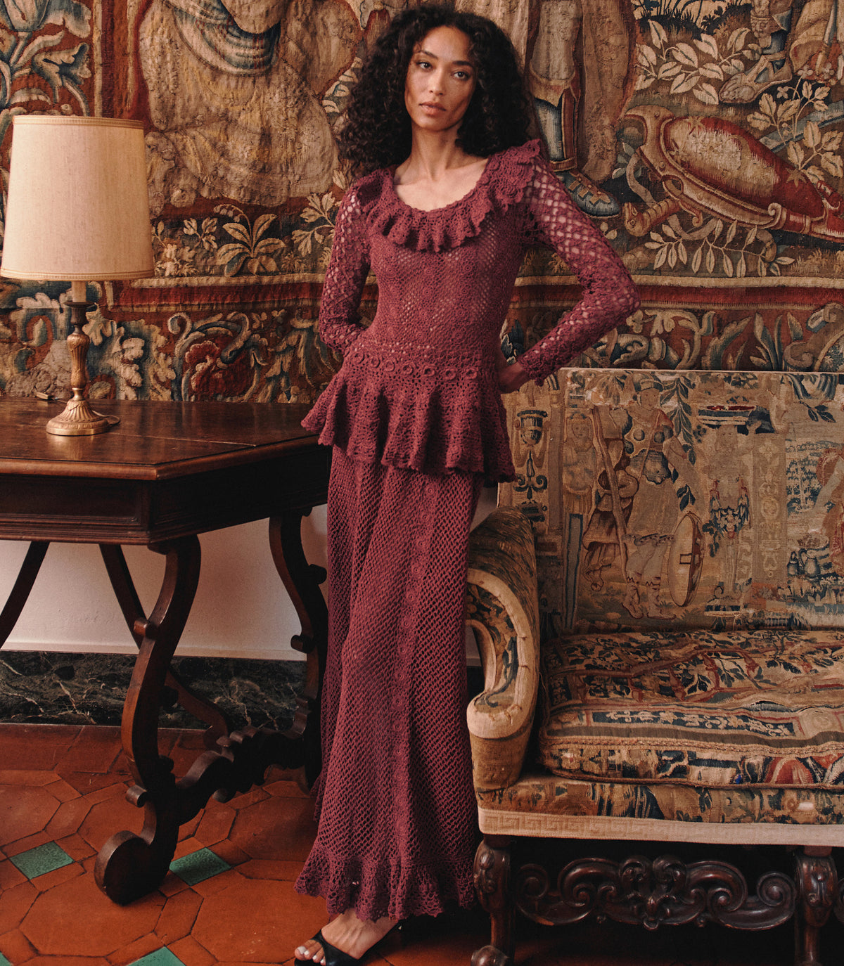 A woman models a long-sleeve maxi dress set in a textured, ruffled burgundy lace. The setting is a vintage room with a tapestry wall, a wooden side table with a lamp, and an ornate armchair.