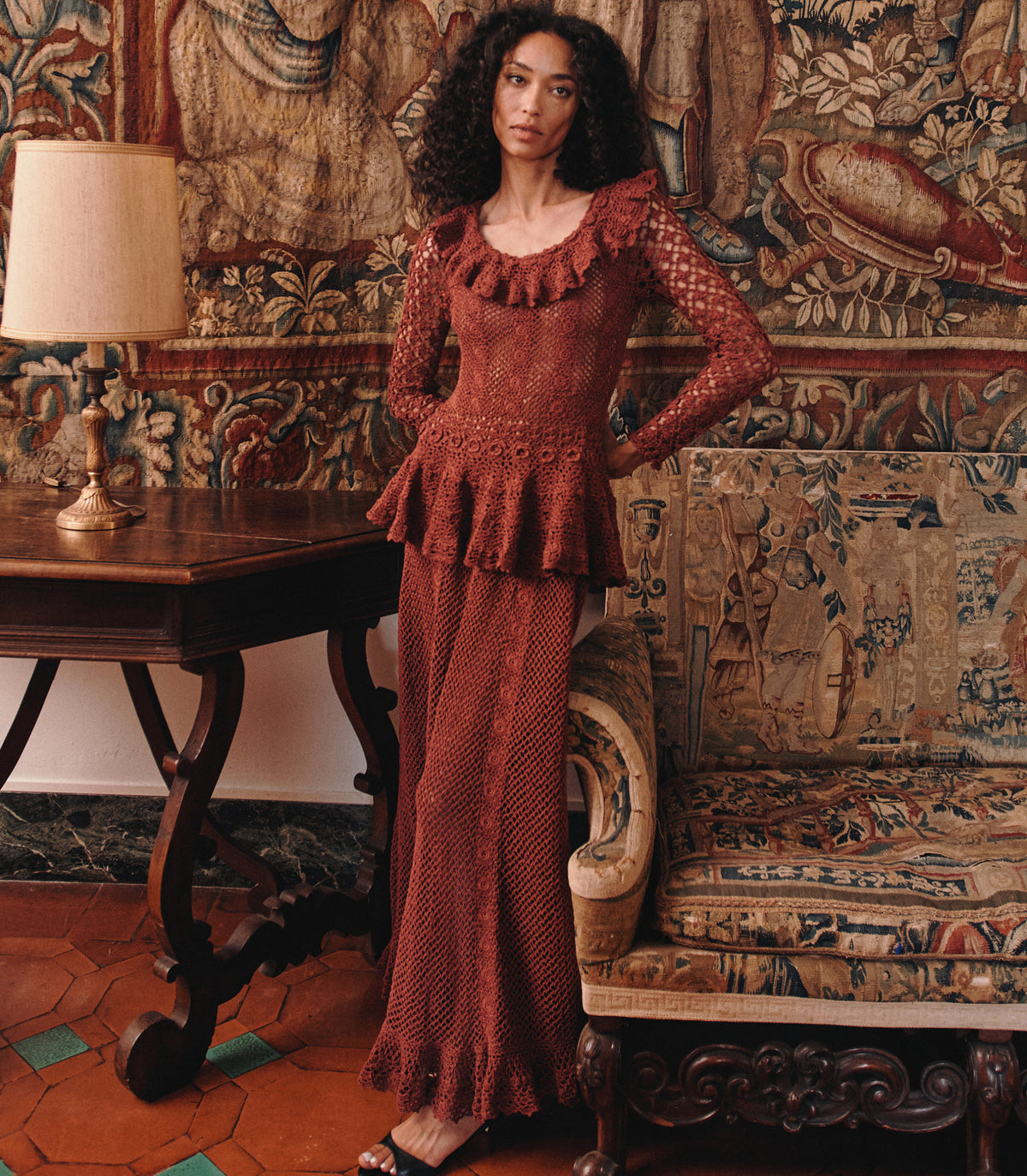 A woman models a rust-brown long-sleeve knit dress with a ruffled neckline and peplum waist, standing in an ornate room with tapestries, a wooden table, and a lamp.