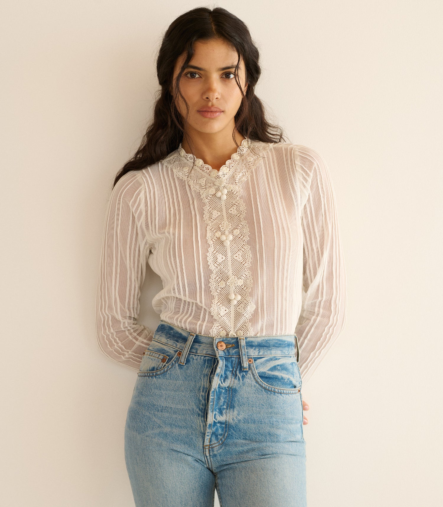 Woman wearing a sheer white blouse with lace details and blue jeans against a plain background