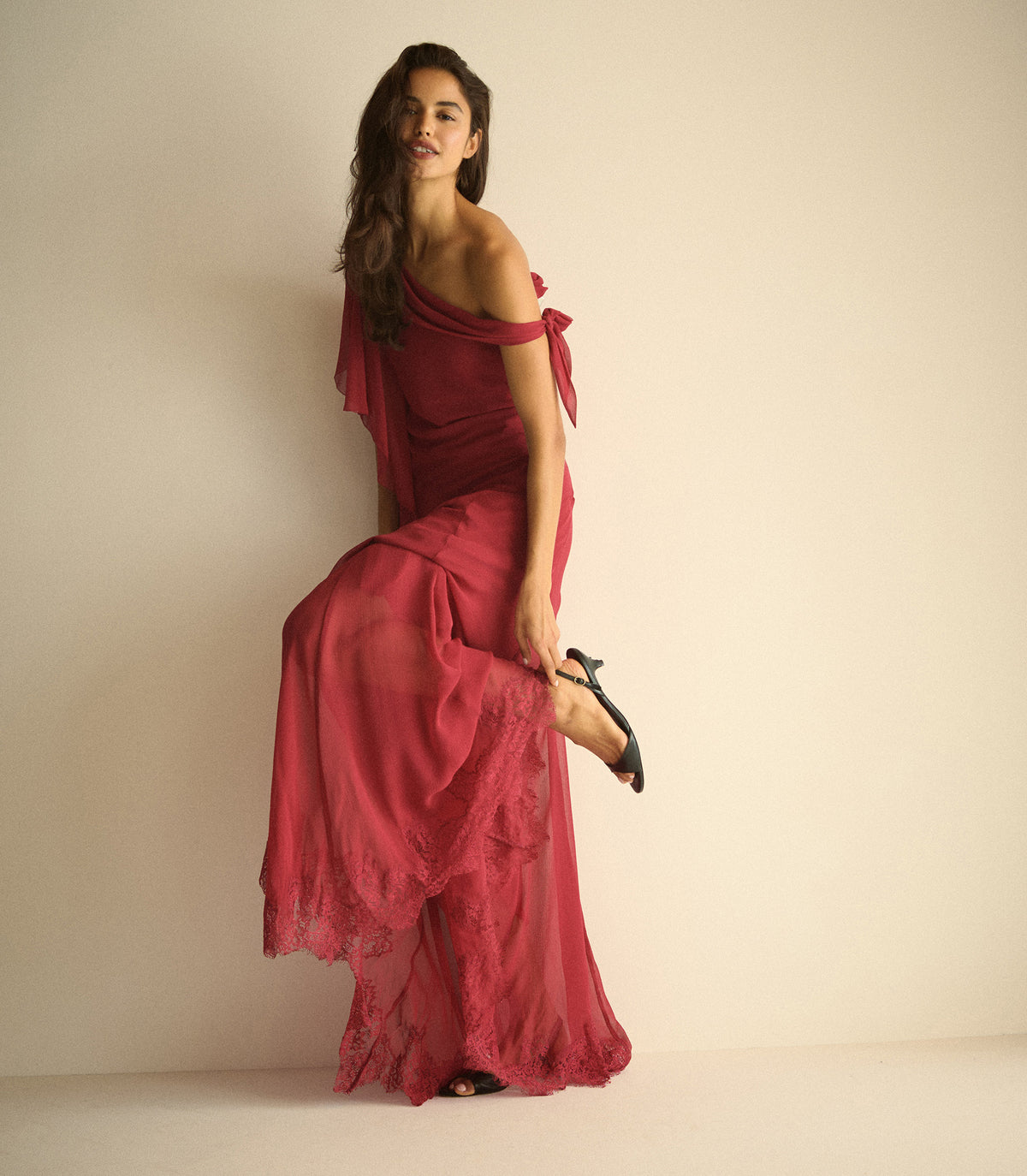 A woman modeling a long, flowing off-the-shoulder dress in deep red with a sheer draped overlay and lace hem, posed against a plain beige studio background. 1
