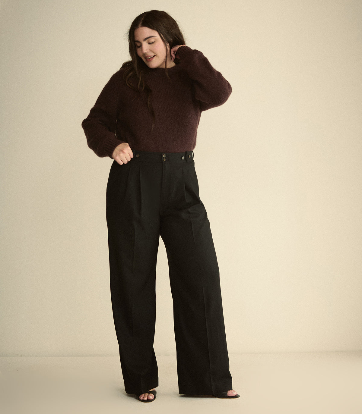A woman modeling a dark brown knit sweater and black wide-leg trousers against a plain beige studio background. 1
