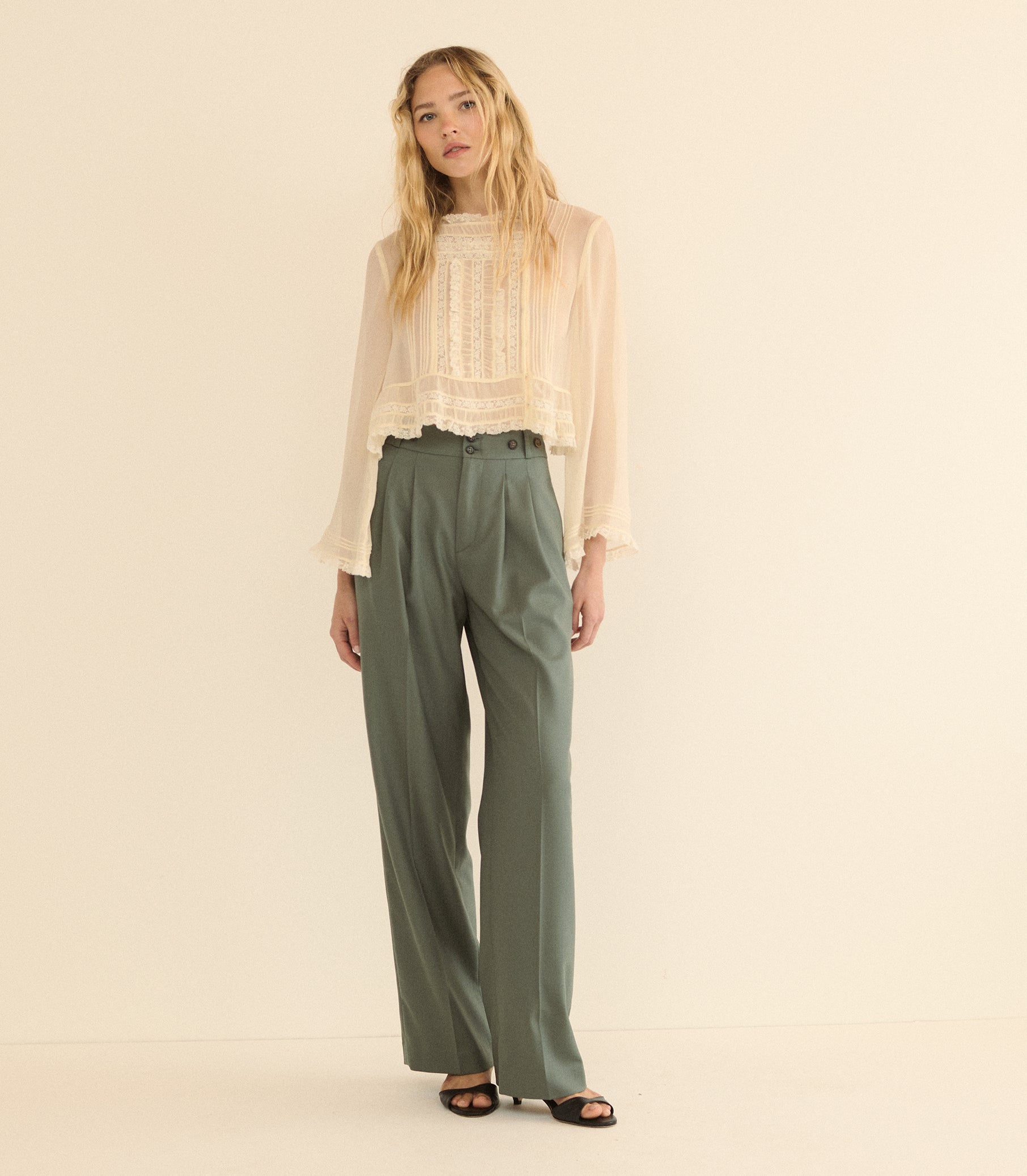 A woman stands against a plain warm cream studio background wearing a sheer long-sleeve cream blouse with a lace front and high-waisted olive green wide-leg trousers. 1
