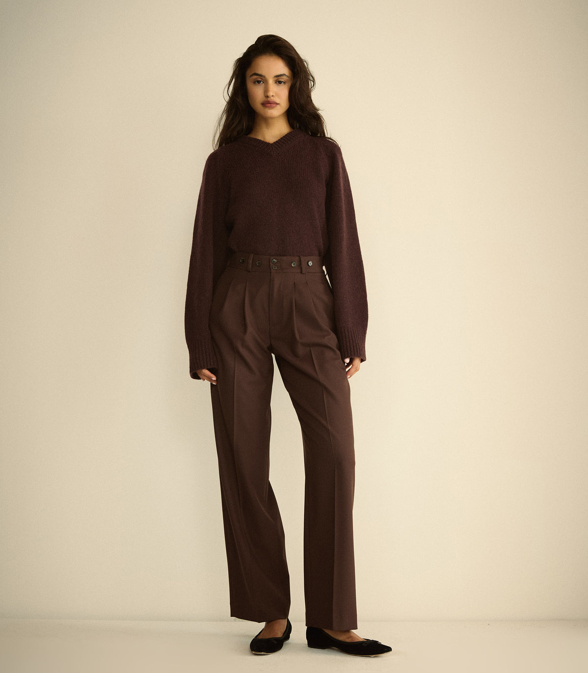 A woman modeling a dark brown long-sleeve knit sweater and matching dark brown wide-leg trousers with a belt, paired with black flats, against a plain beige studio background.