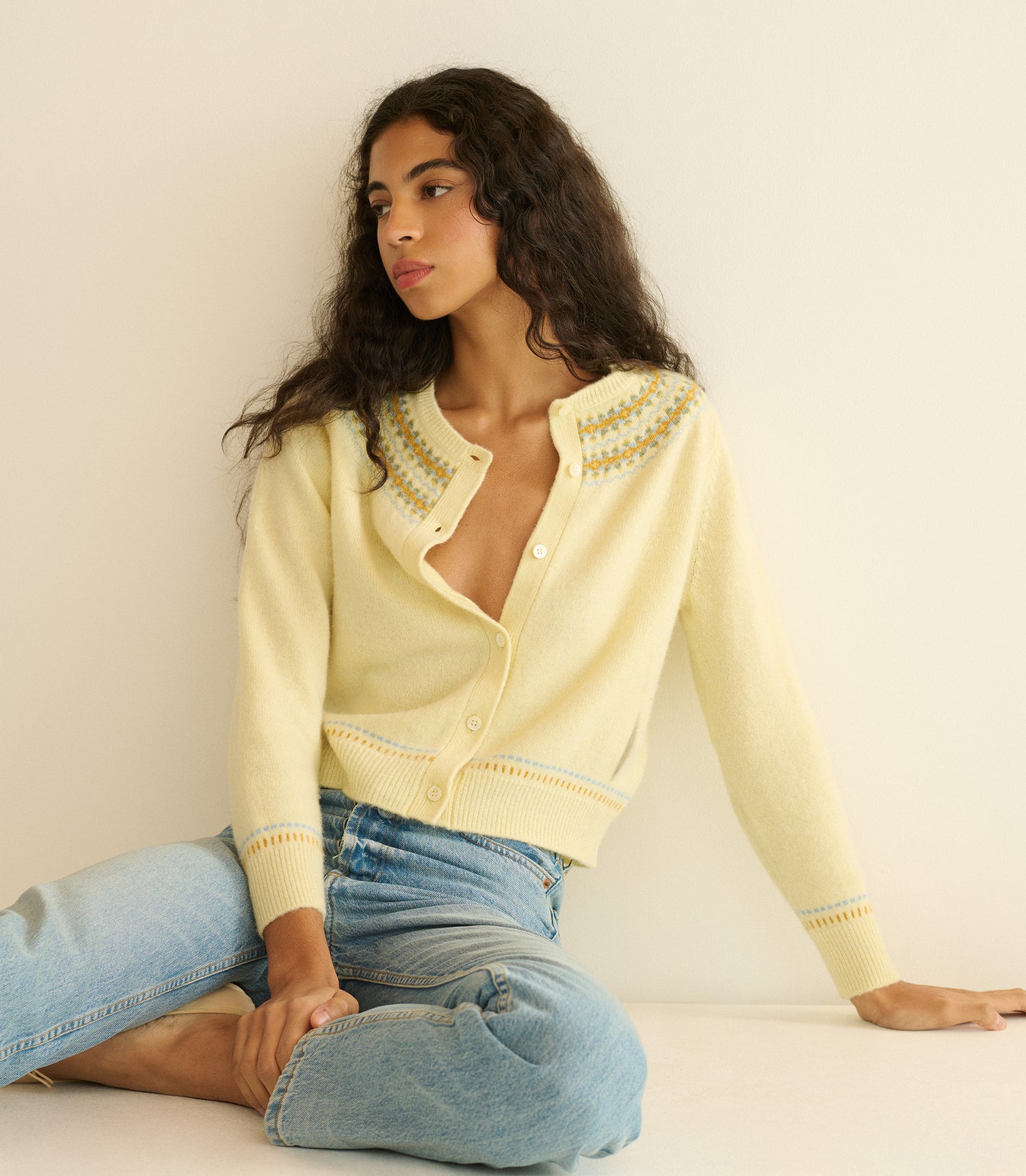 Woman wearing a light yellow cardigan with intricate patterns, sitting against a plain background.