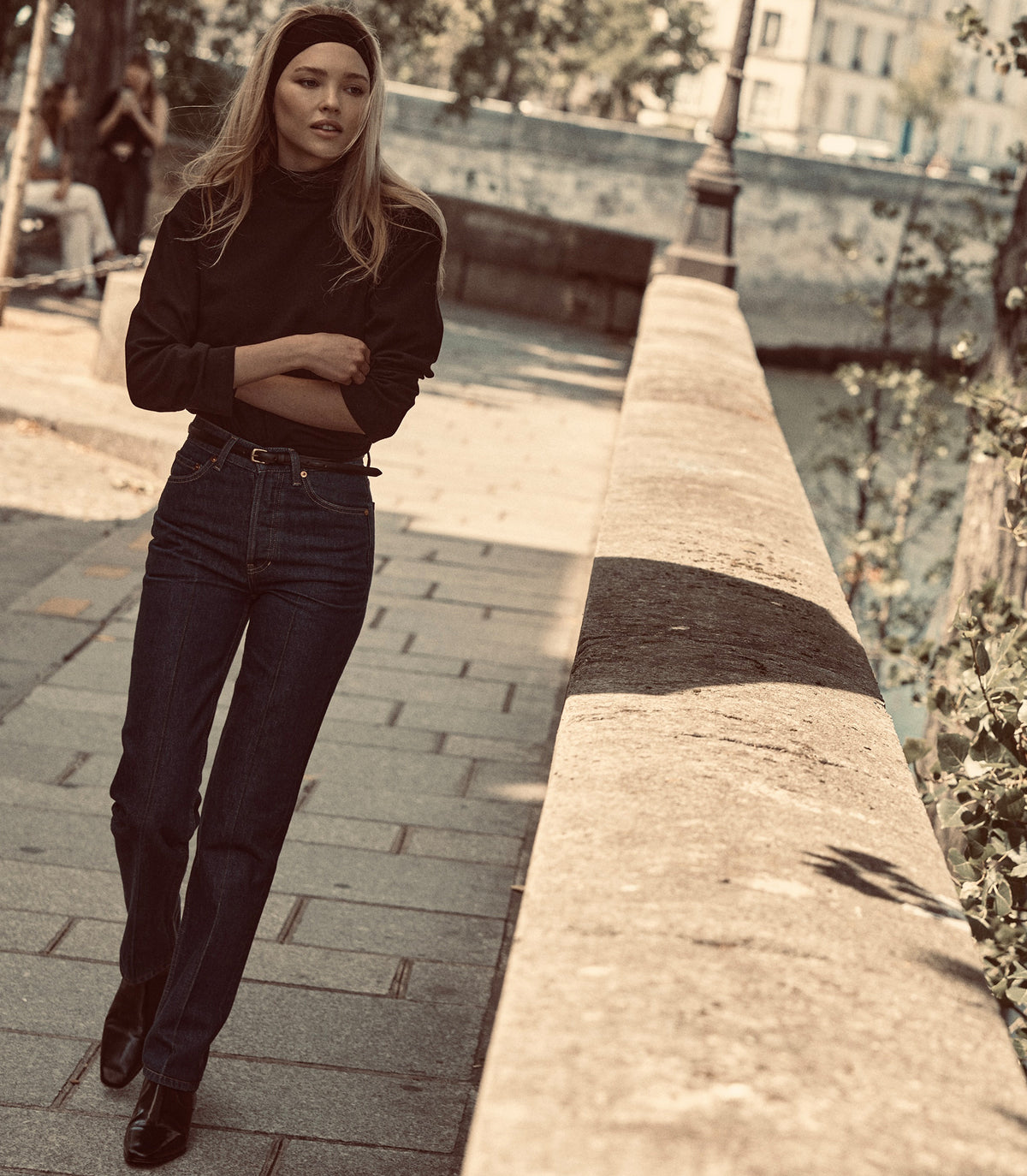 A woman wearing a black long-sleeve mock-neck top, dark blue jeans, and a black headband walks outdoors.