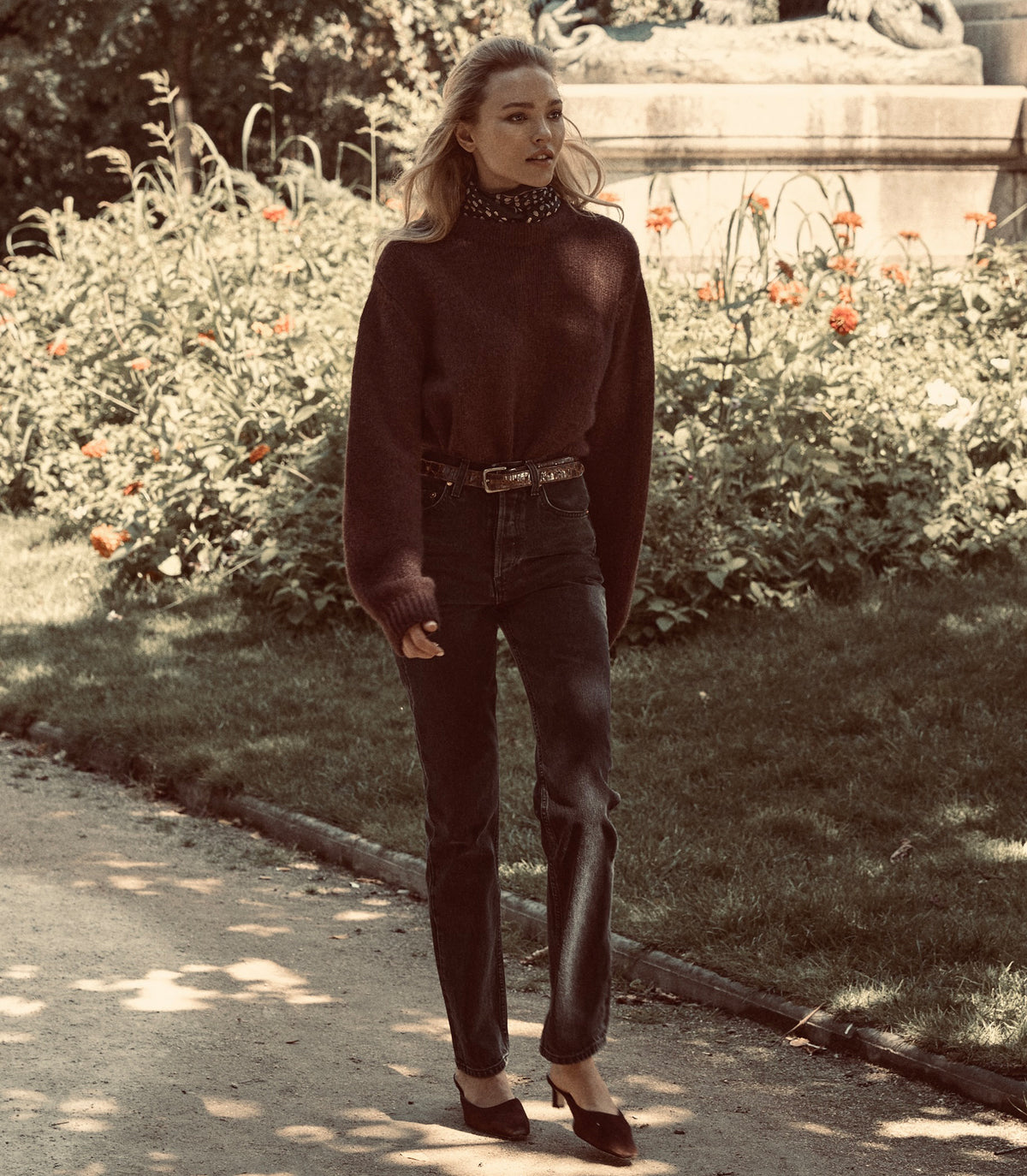 A woman stands outdoors in a park-like setting wearing a dark brown ribbed mock-neck sweater, dark wash jeans, and dark pointed-toe heels. 7
