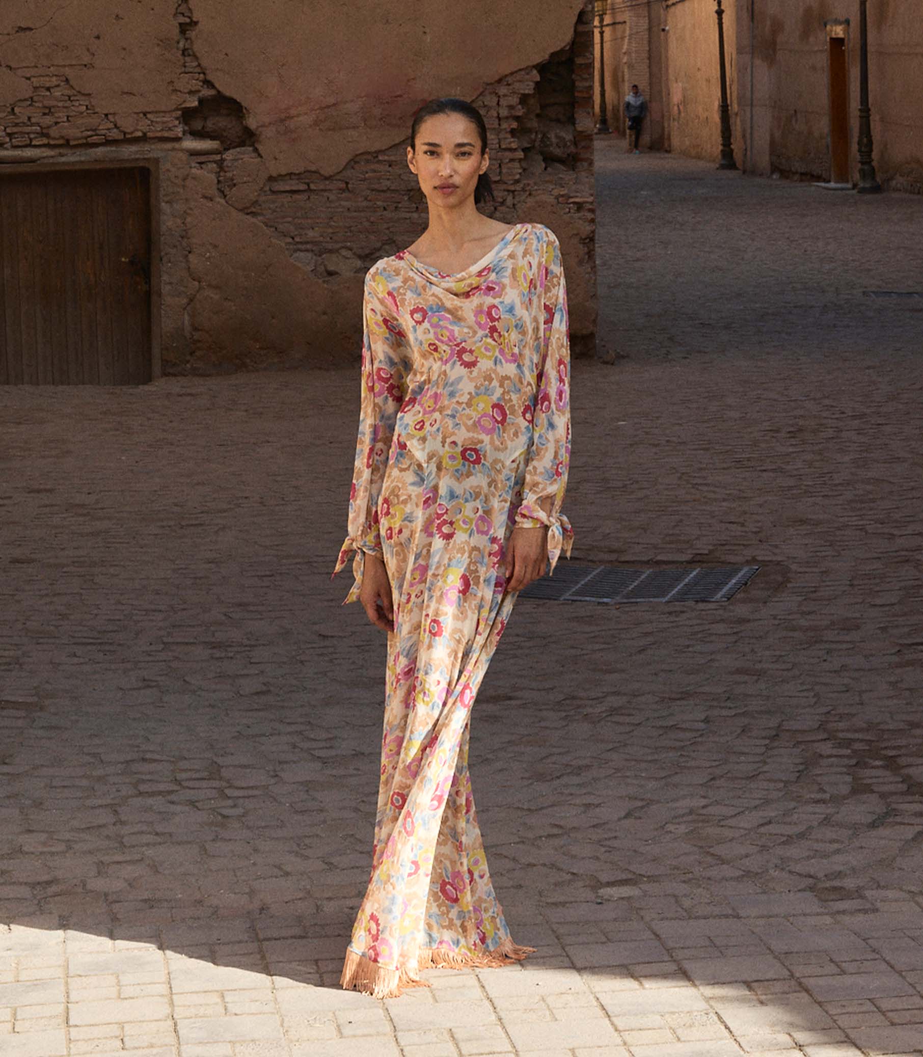 Woman in a long floral dress standing in a sunlit alleyway.
