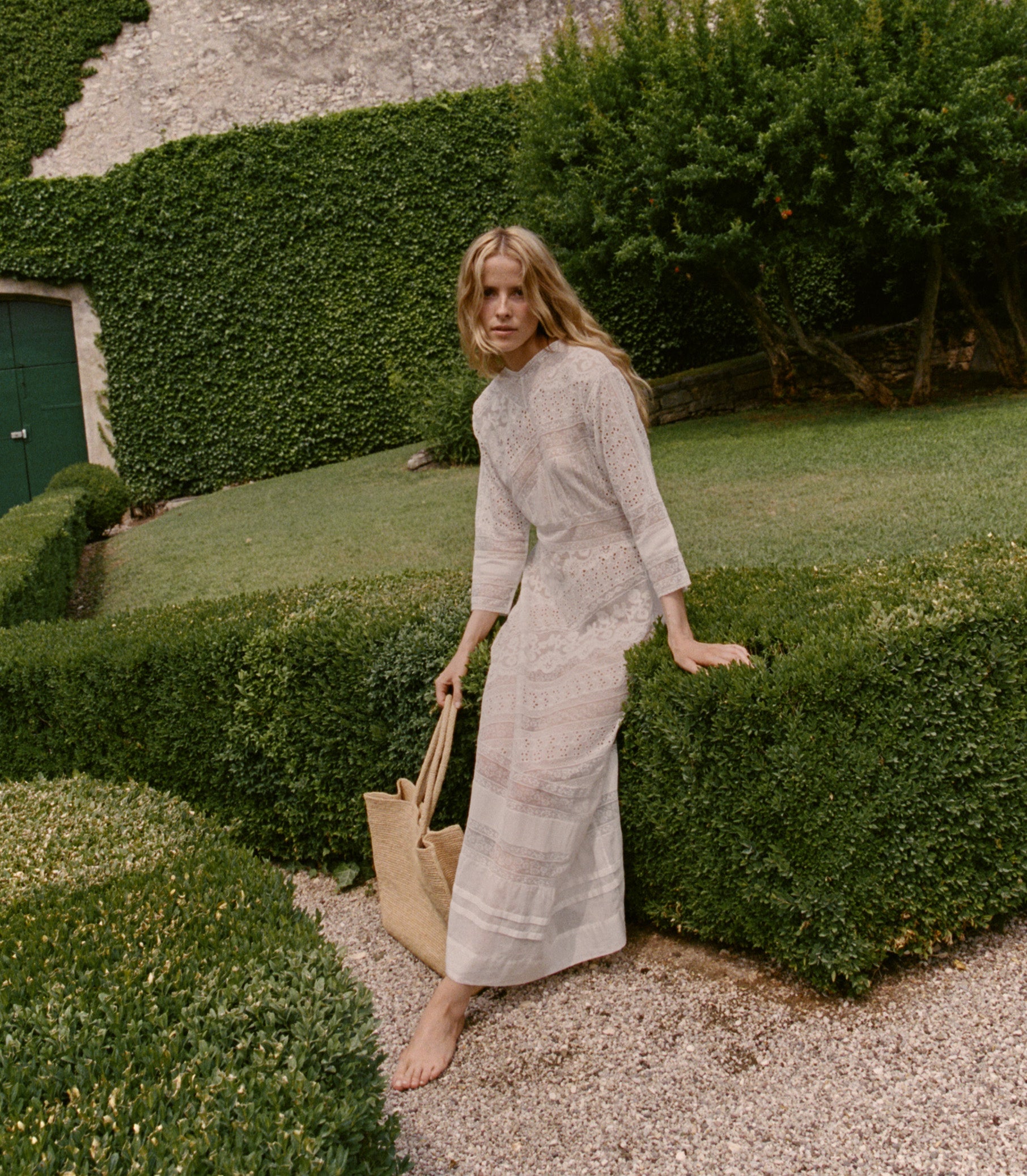 Woman in a light dress standing next to a well-manicured hedge in a garden.