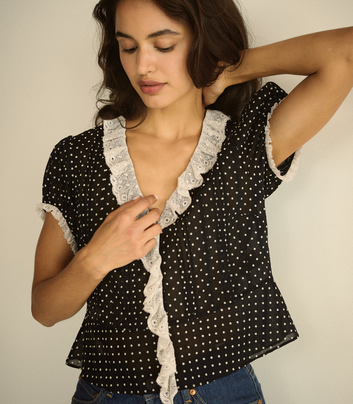 A woman wearing a black short-sleeve blouse with white polka dots and a white lace ruffle trim along the front and cuffs, paired with blue jeans against a plain beige background.