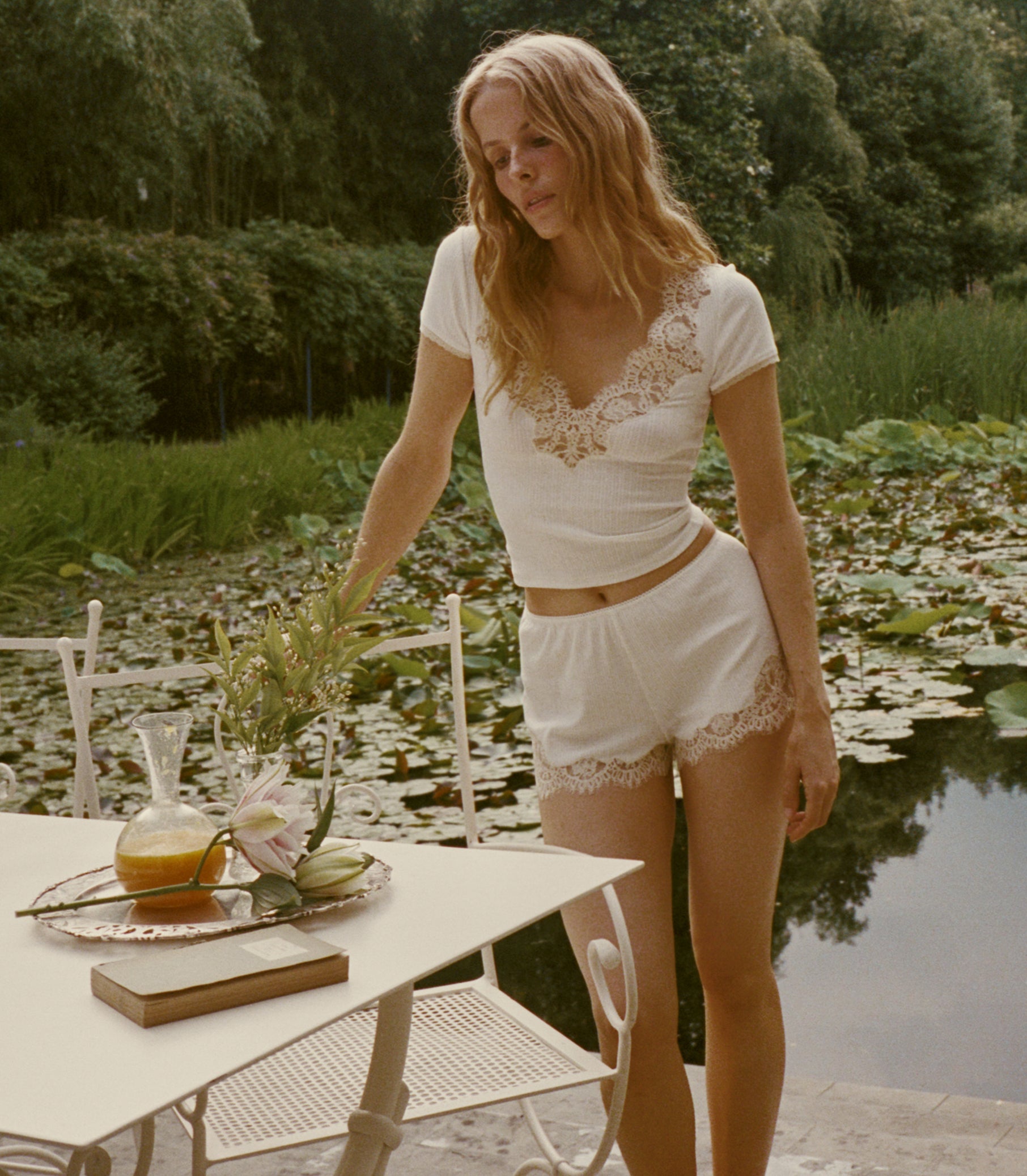 A woman wearing a white two-piece ribbed set with lace trim stands beside a white outdoor table by a pond with lily pads and trees in the background.
