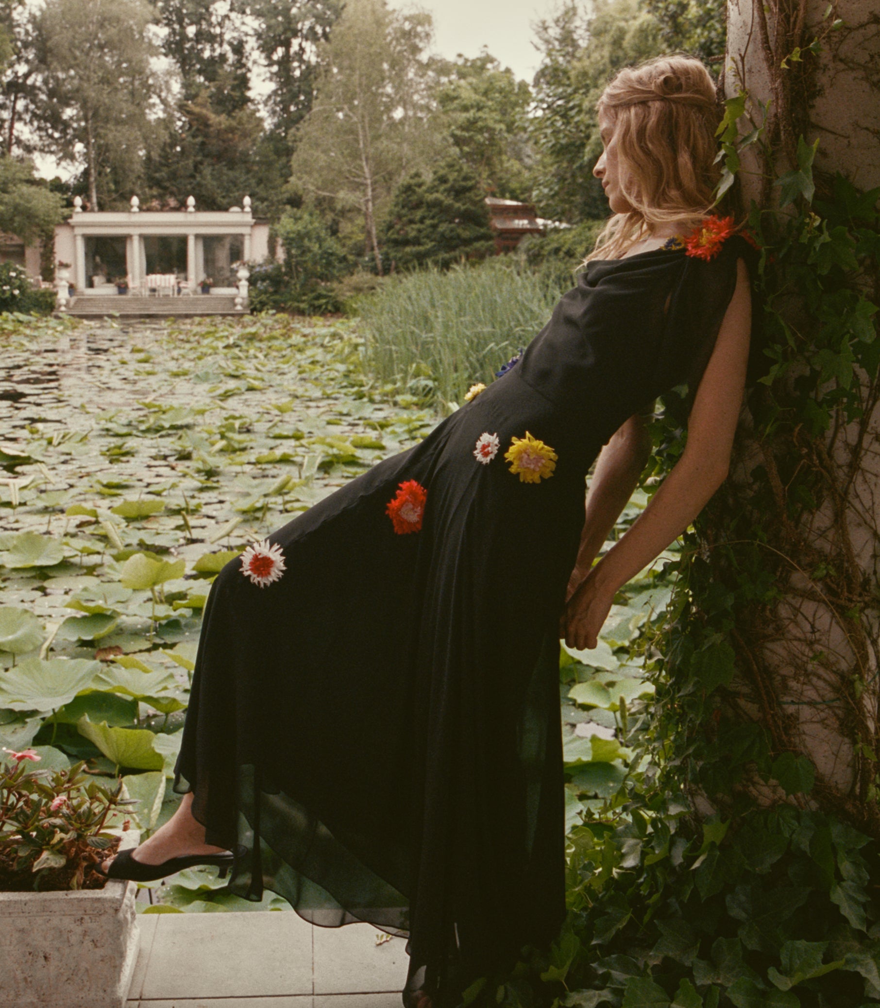 A woman in a black maxi dress with colorful flower appliqués leans against a vine-covered wall beside a lily pond. 1
