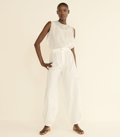 A model wearing a white top with a lace-trimmed round neckline and white wide-leg pants, paired with brown leather sandals, against a solid cream background.