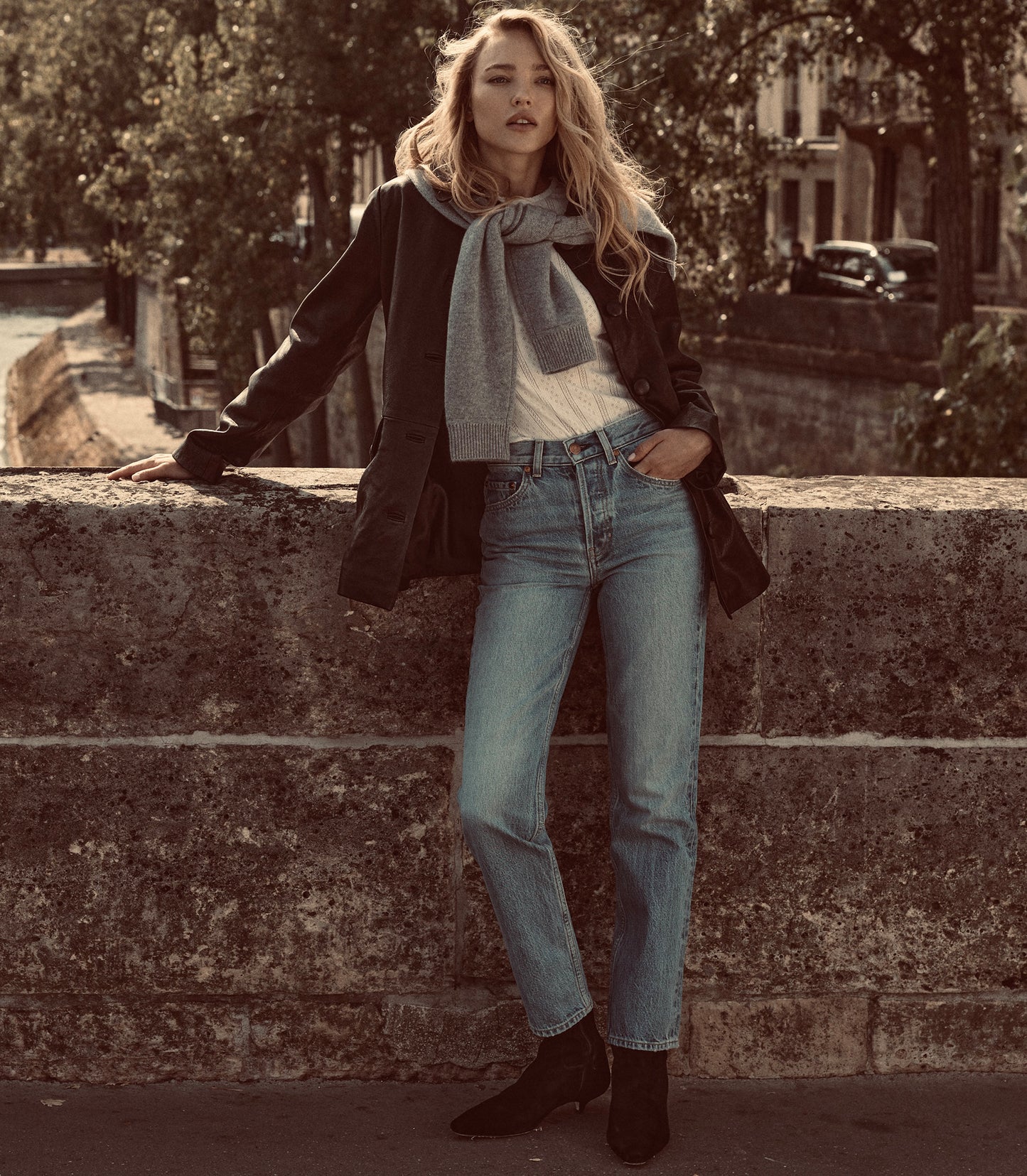 A woman stands outdoors leaning against a stone wall, wearing a black coat, light gray scarf, white top, light blue jeans, and black ankle boots.