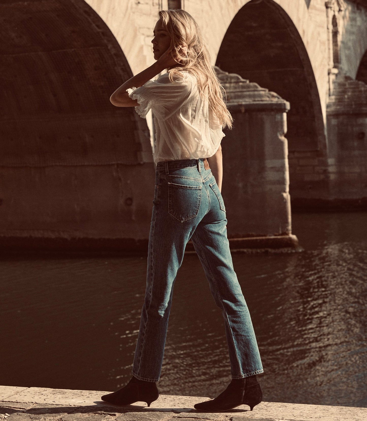 A woman stands by a river under a stone bridge, wearing high-waisted blue jeans, a white blouse, and black ankle boots.