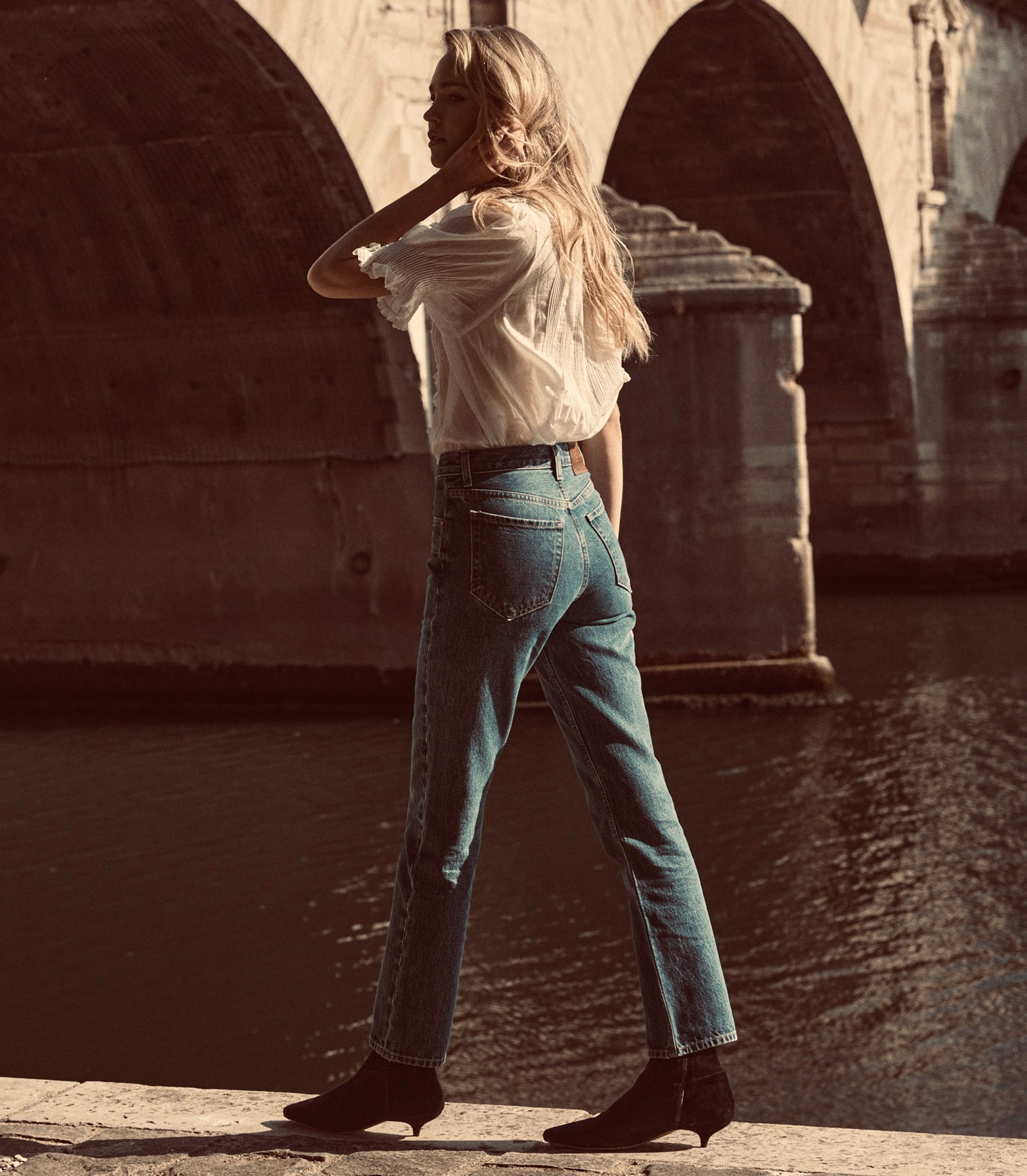 A woman stands by a river under a stone bridge, wearing high-waisted blue jeans, a white blouse, and black ankle boots. 1
