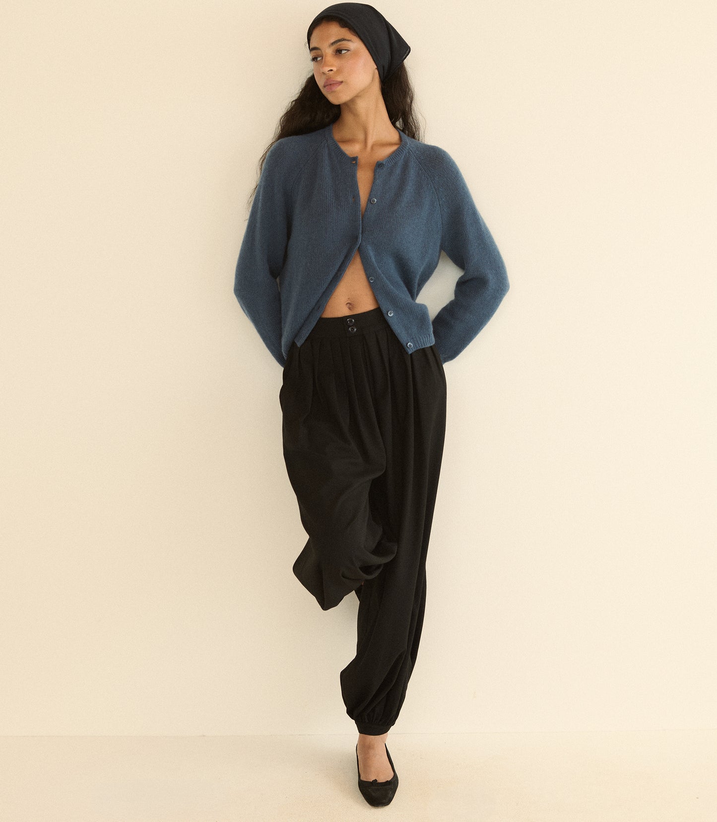 A woman modeling a blue cropped cardigan, black harem pants, a black beanie, and black flats against a plain cream background.