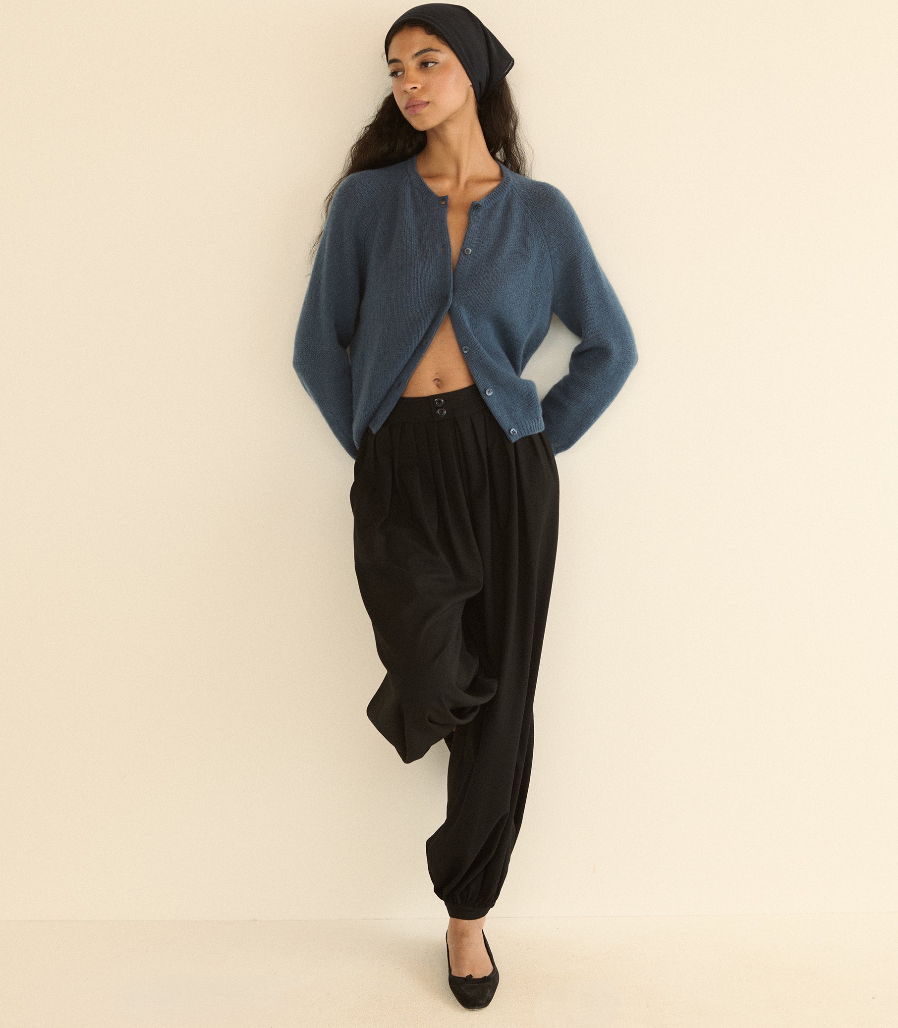 A woman modeling a blue cropped cardigan, black harem pants, a black beanie, and black flats against a plain cream background. 1

