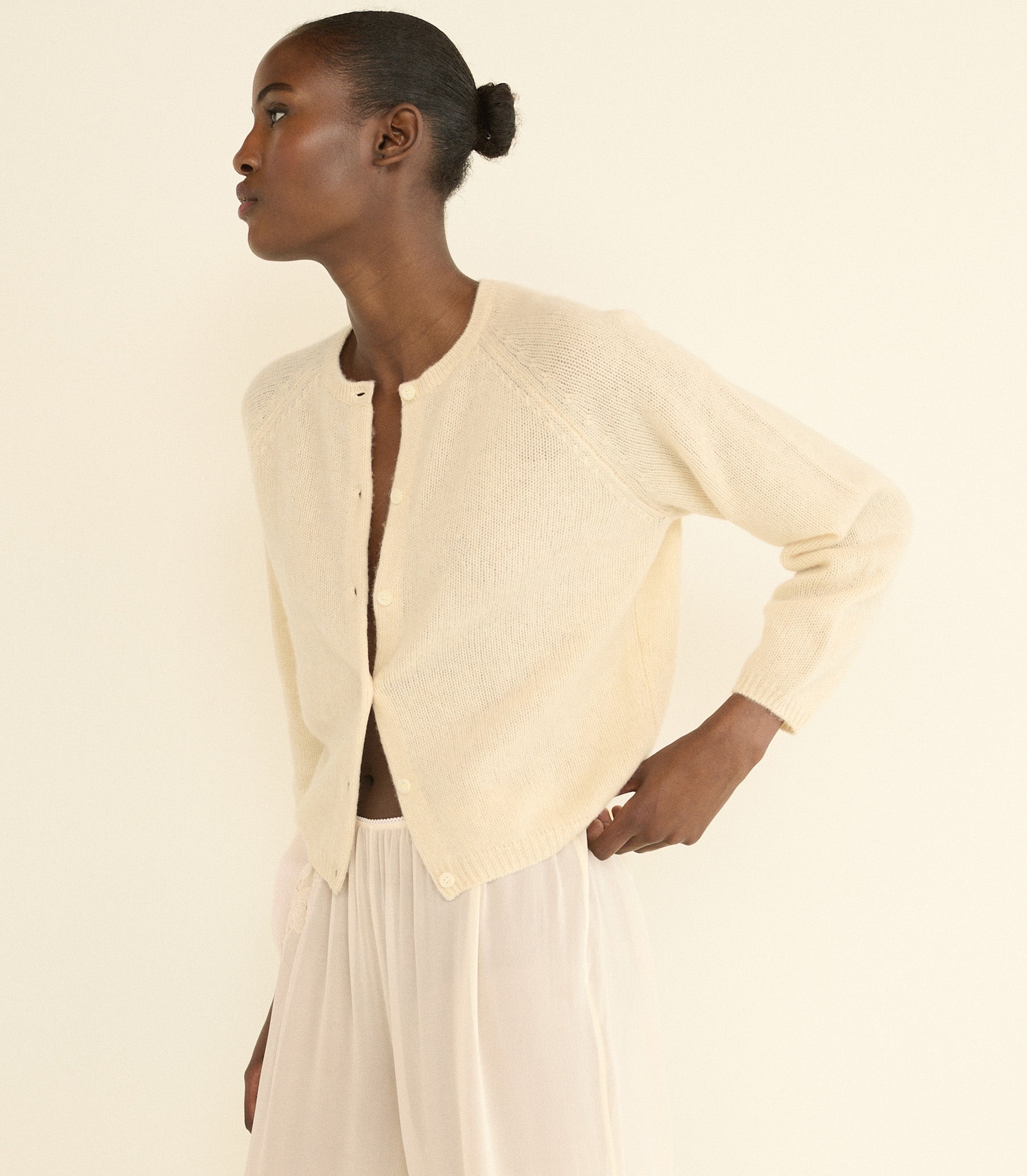 A model wearing a cream-colored, long-sleeve knit cardigan and cream trousers, shown from a side profile against a solid cream background. 1
