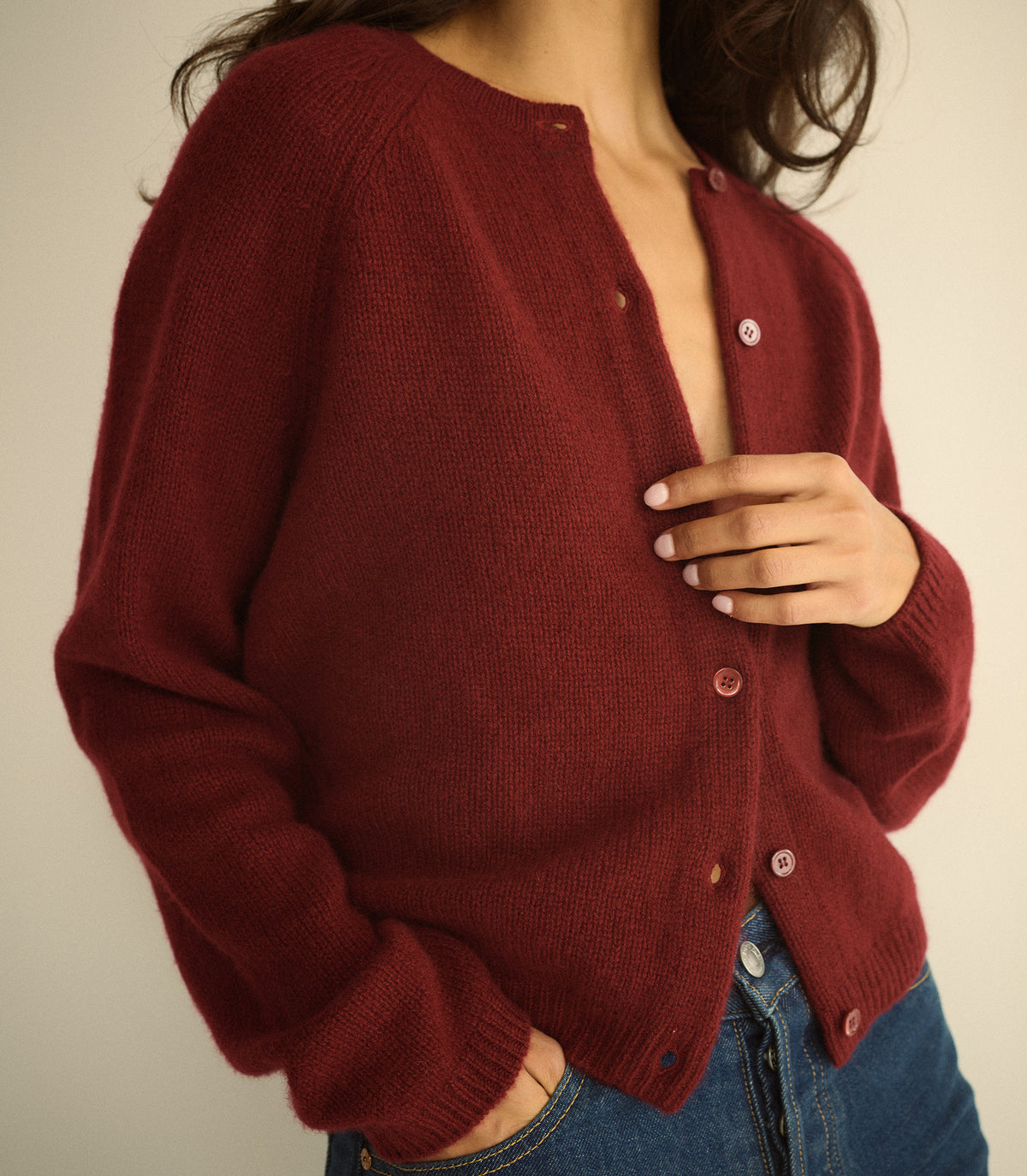 A woman wearing a burgundy knit cardigan sweater with buttons, paired with blue jeans, shown from the chest down against a plain light beige background. 1
