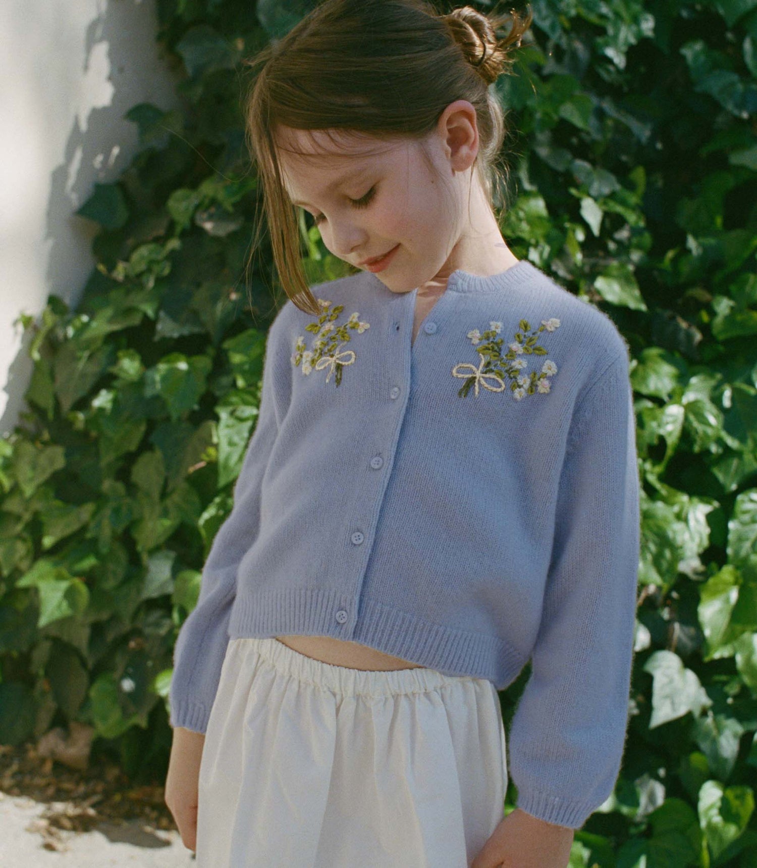 Young girl wearing a light blue cardigan with floral embroidery against a green leafy background
