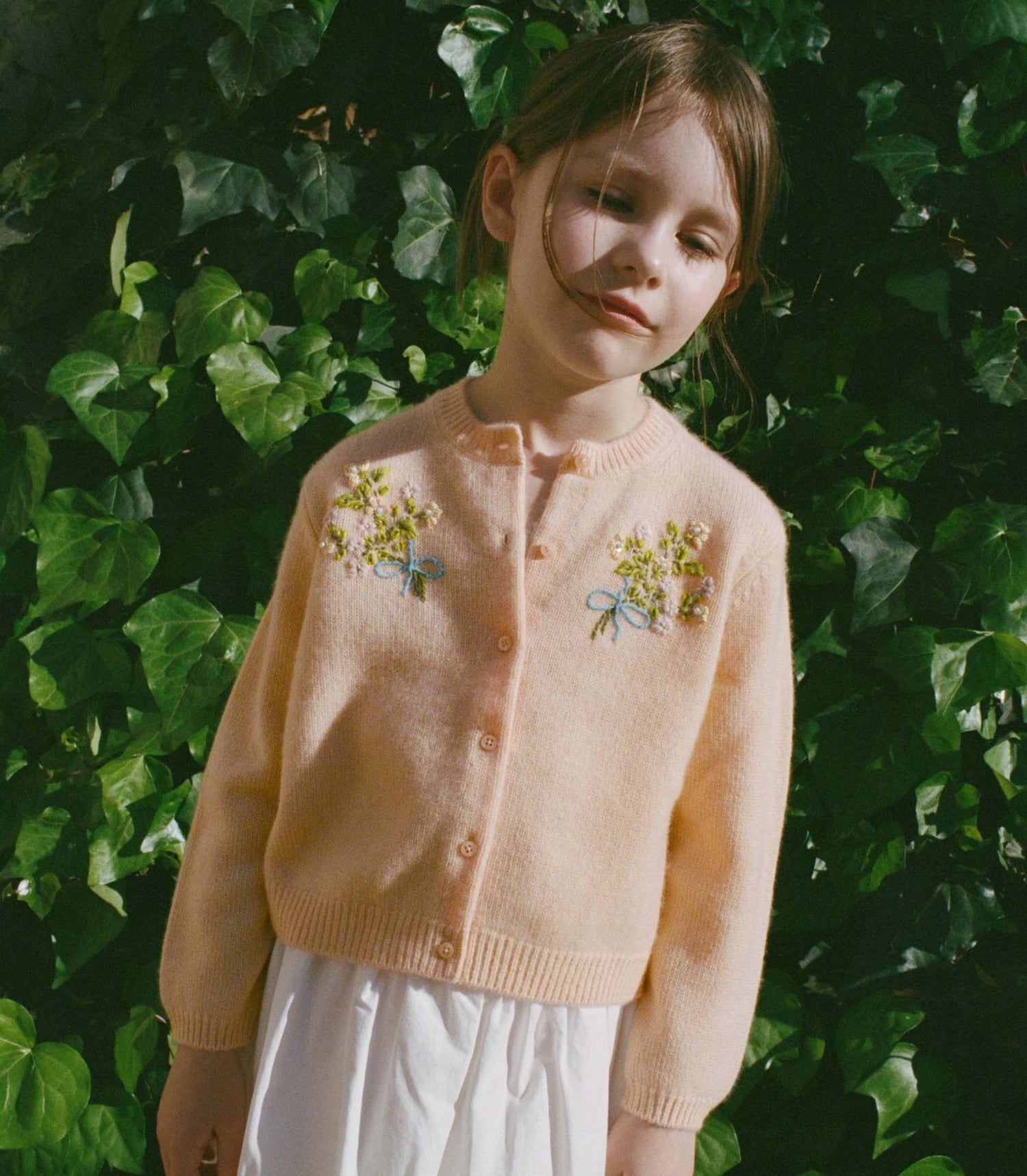 Young girl wearing a light pink cardigan with floral embroidery against a green leafy background