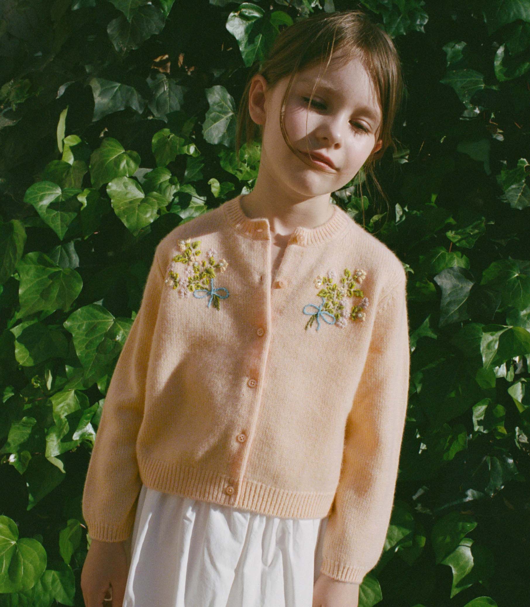 Young girl wearing a light pink cardigan with floral embroidery against a green leafy background 1
