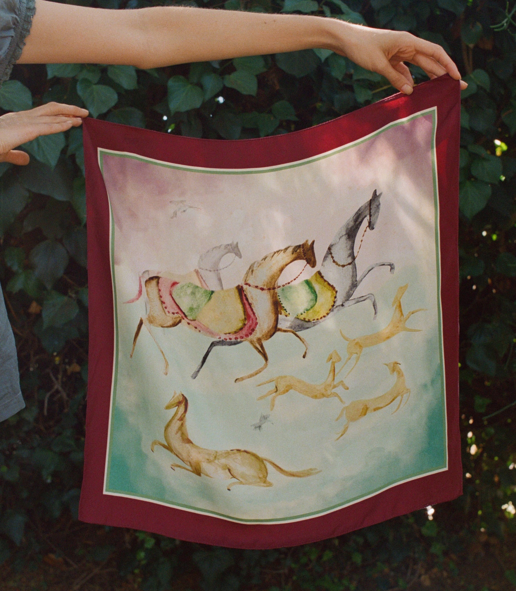 A square silk bandana with a deep red border and a central illustration of horses and gazelles, held up by a person's hands against a background of green foliage. 1

