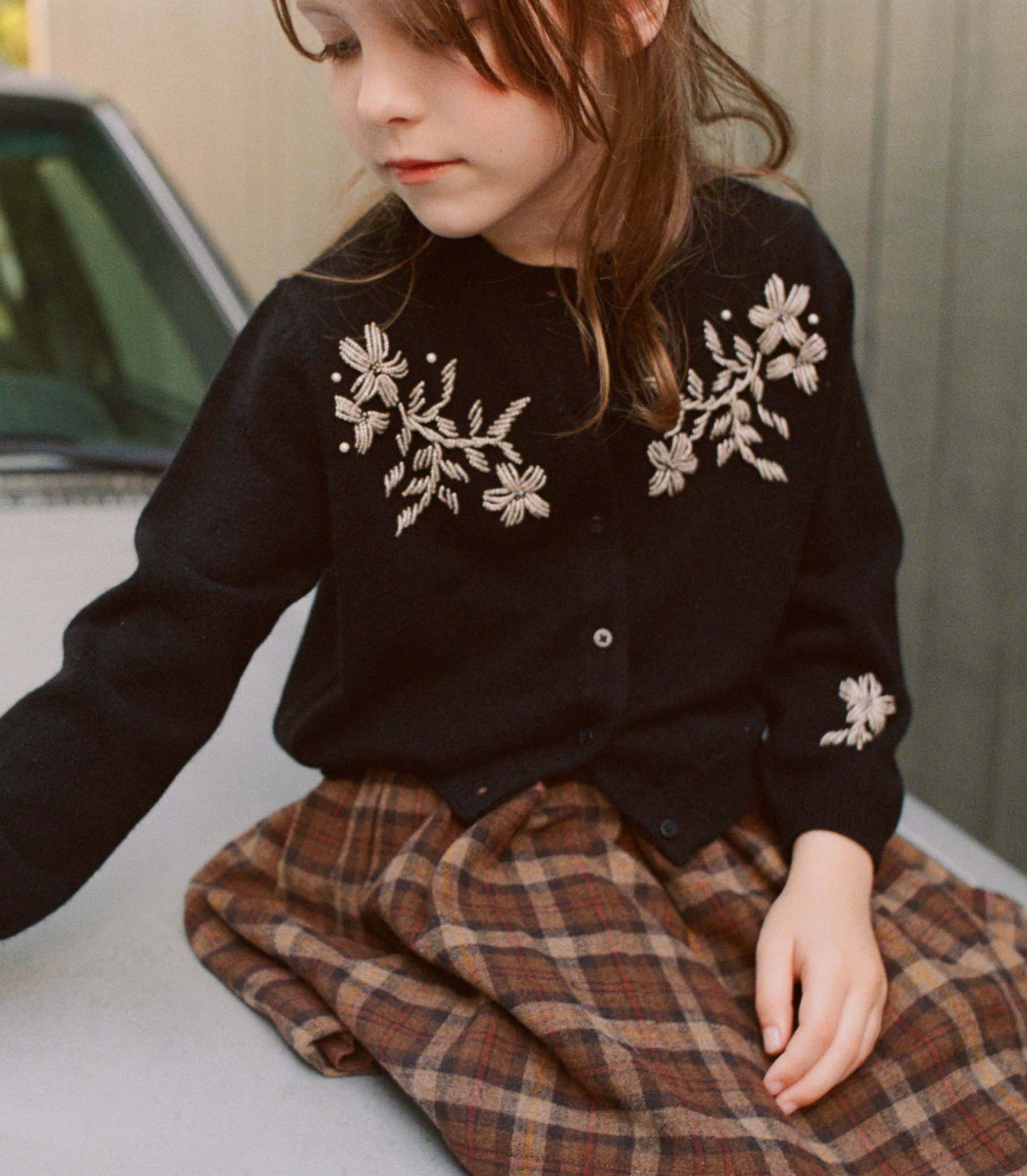 Child wearing a black cardigan with floral embroidery and a plaid skirt sitting on a car. 1