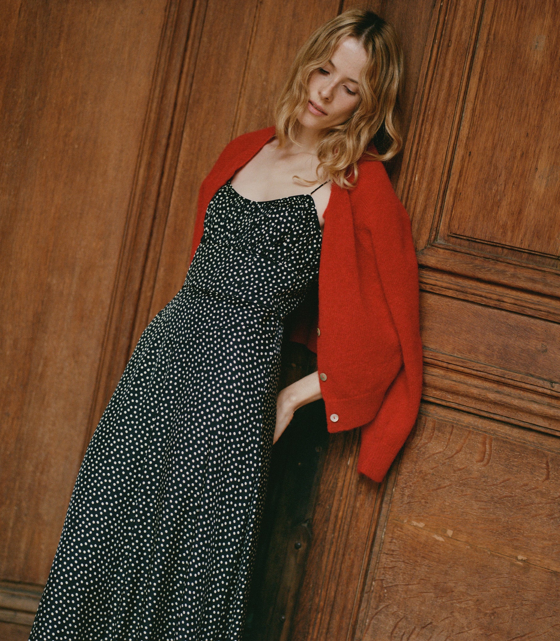 A person wearing a black and white polka dot dress with a red cardigan over her shoulders.