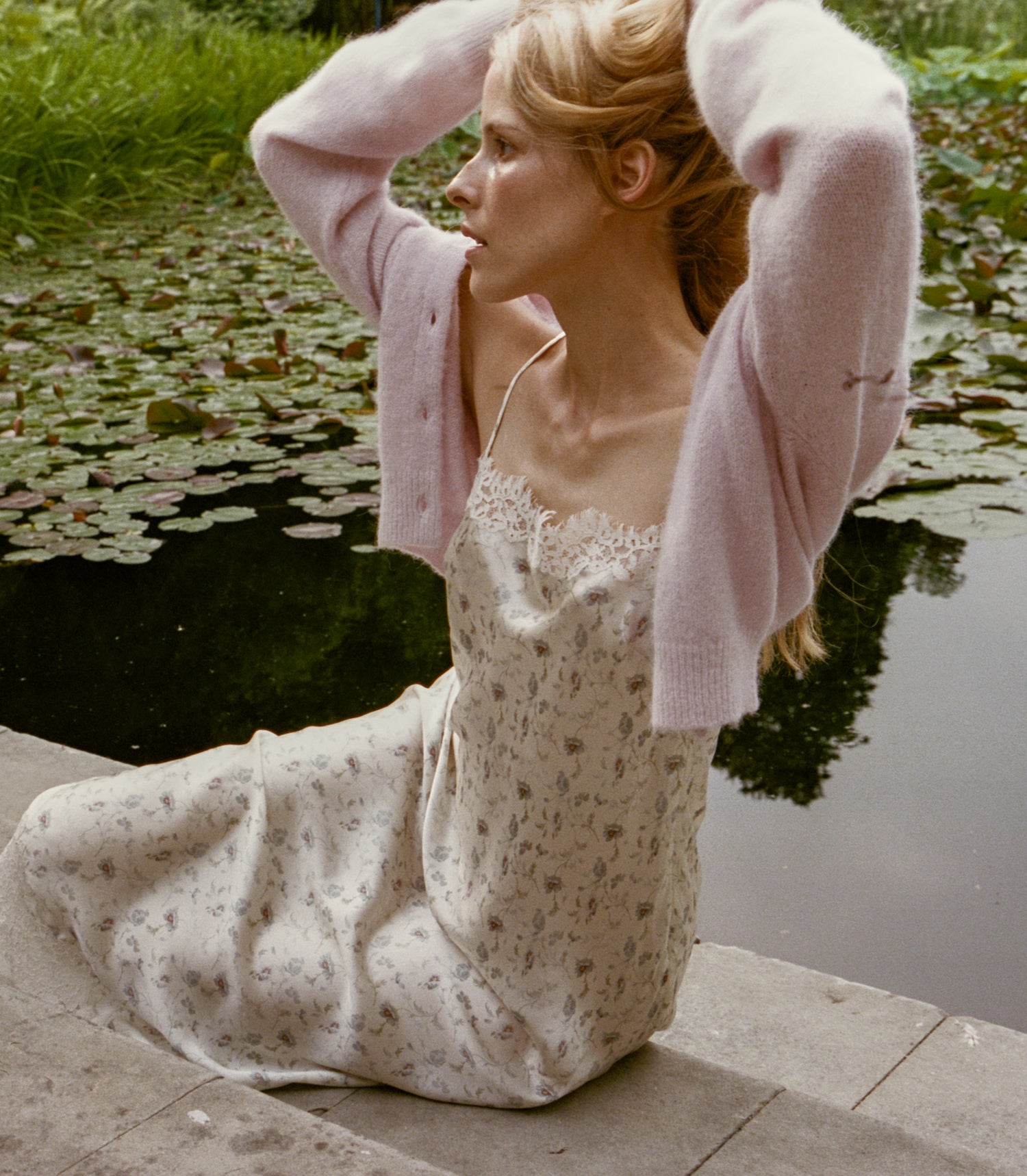 A woman sits by a pond wearing a cream floral slip dress with a light pink knit cardigan draped off her shoulders. She has her hands raised to her head, with lily pads and green grass visible in the background.