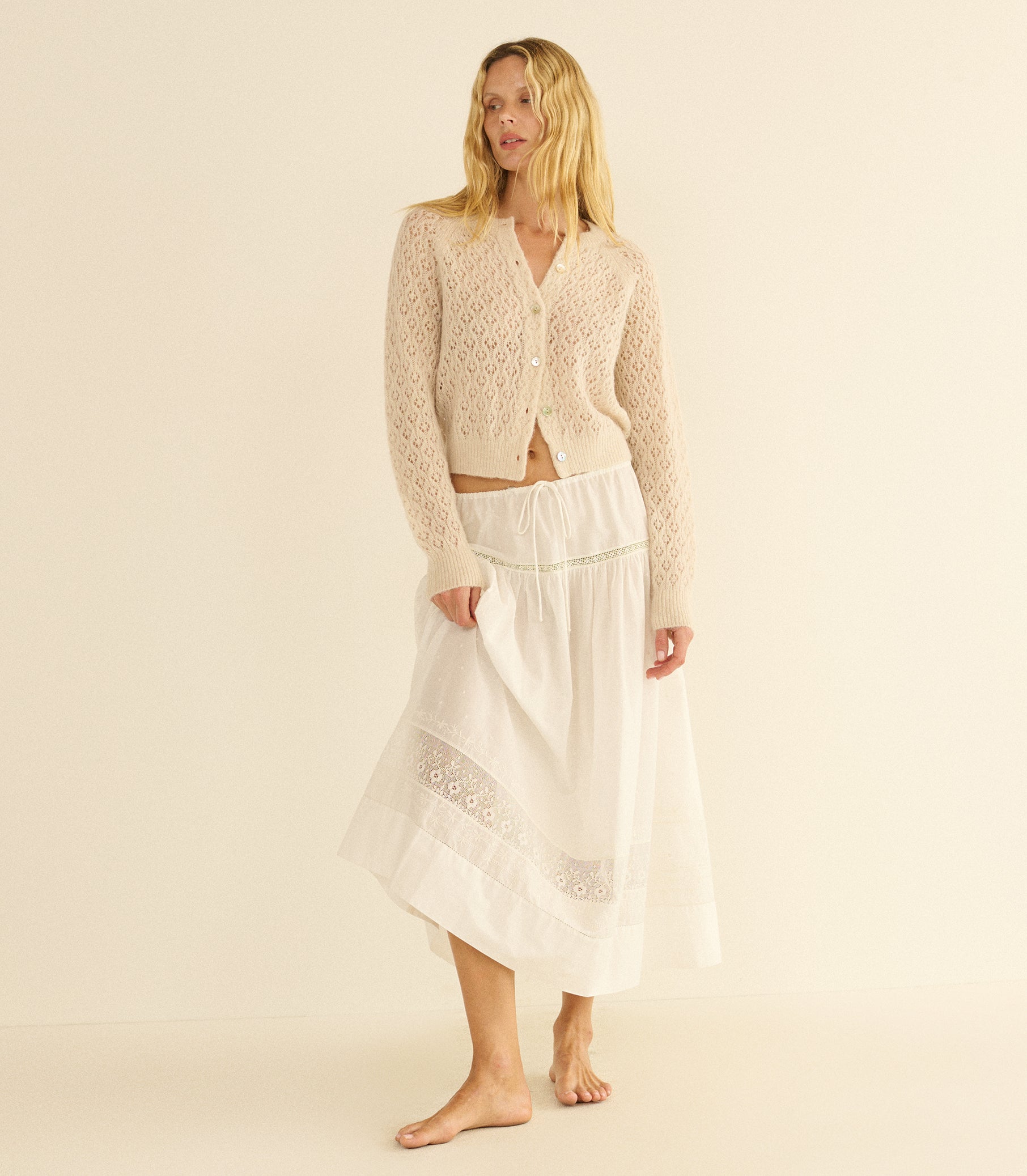 Woman wearing a beige pointelle cardigan and white skirt on a beige background 1
