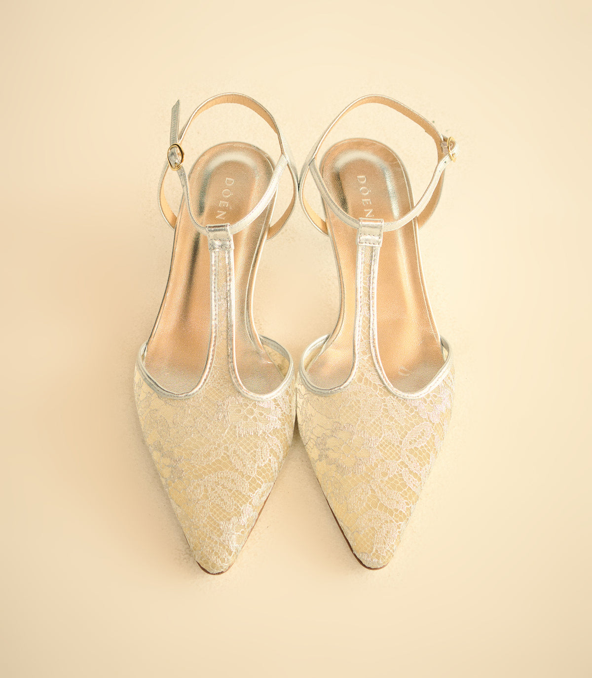 A pair of pointed-toe high heels with ankle straps, featuring a textured champagne-gold lace upper and a thin metallic silver T-strap, shown on a solid light beige background.