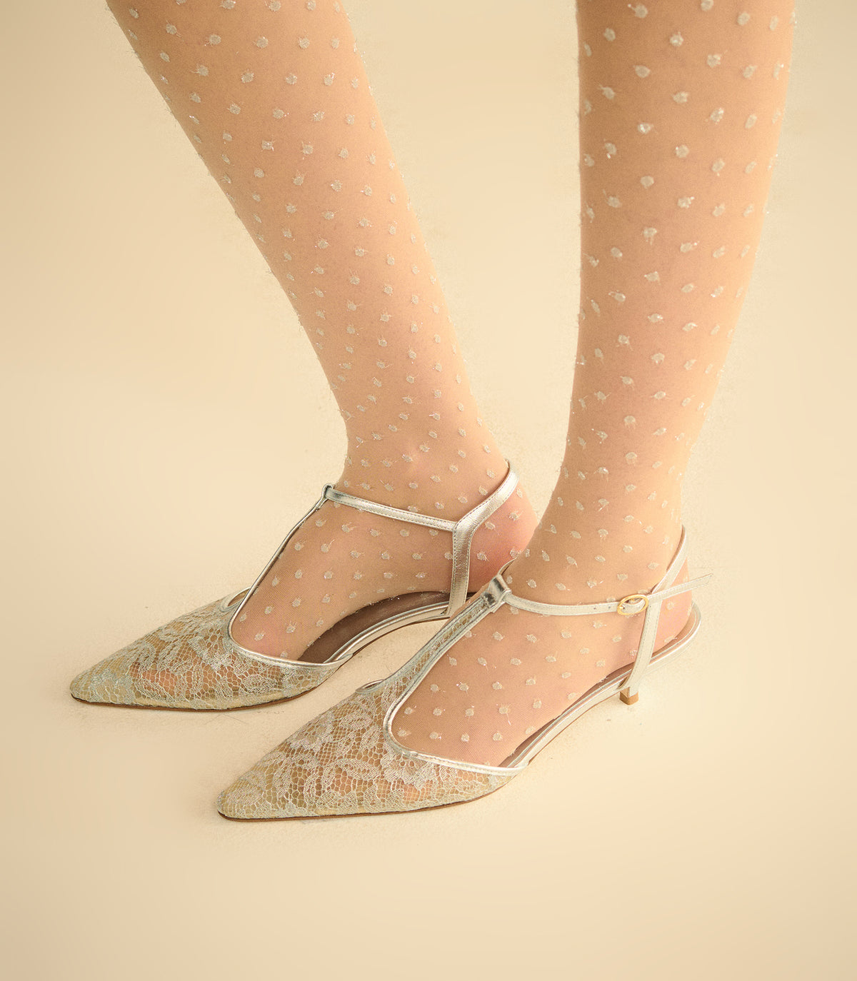 A pair of pointed-toe silver metallic T-strap heels with a subtle floral lace overlay, shown on a model wearing sheer beige tights with small silver dots, against a solid light beige background.