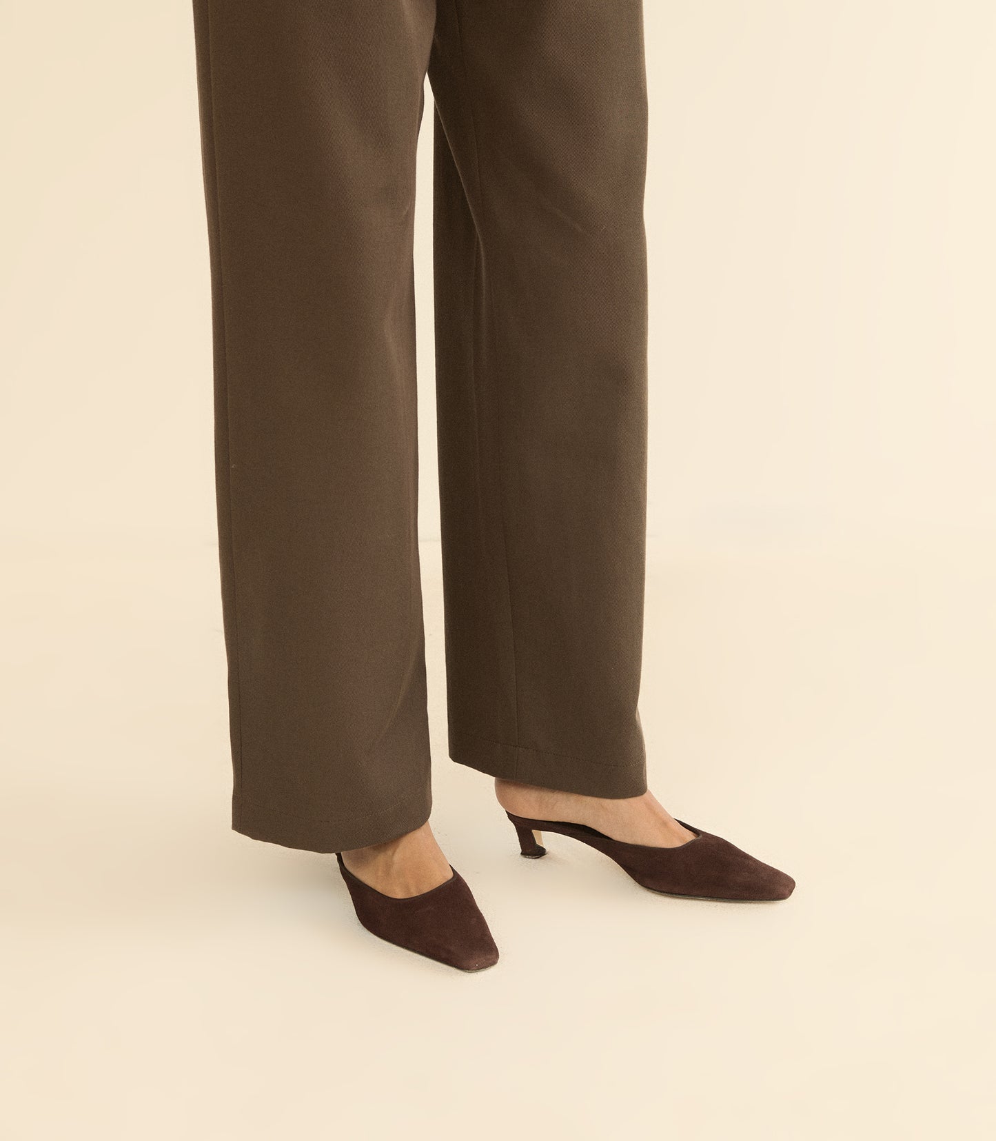 A person wearing dark brown wide-leg trousers and matching pointed-toe mule heels, shown from the knees down against a plain warm beige studio background.