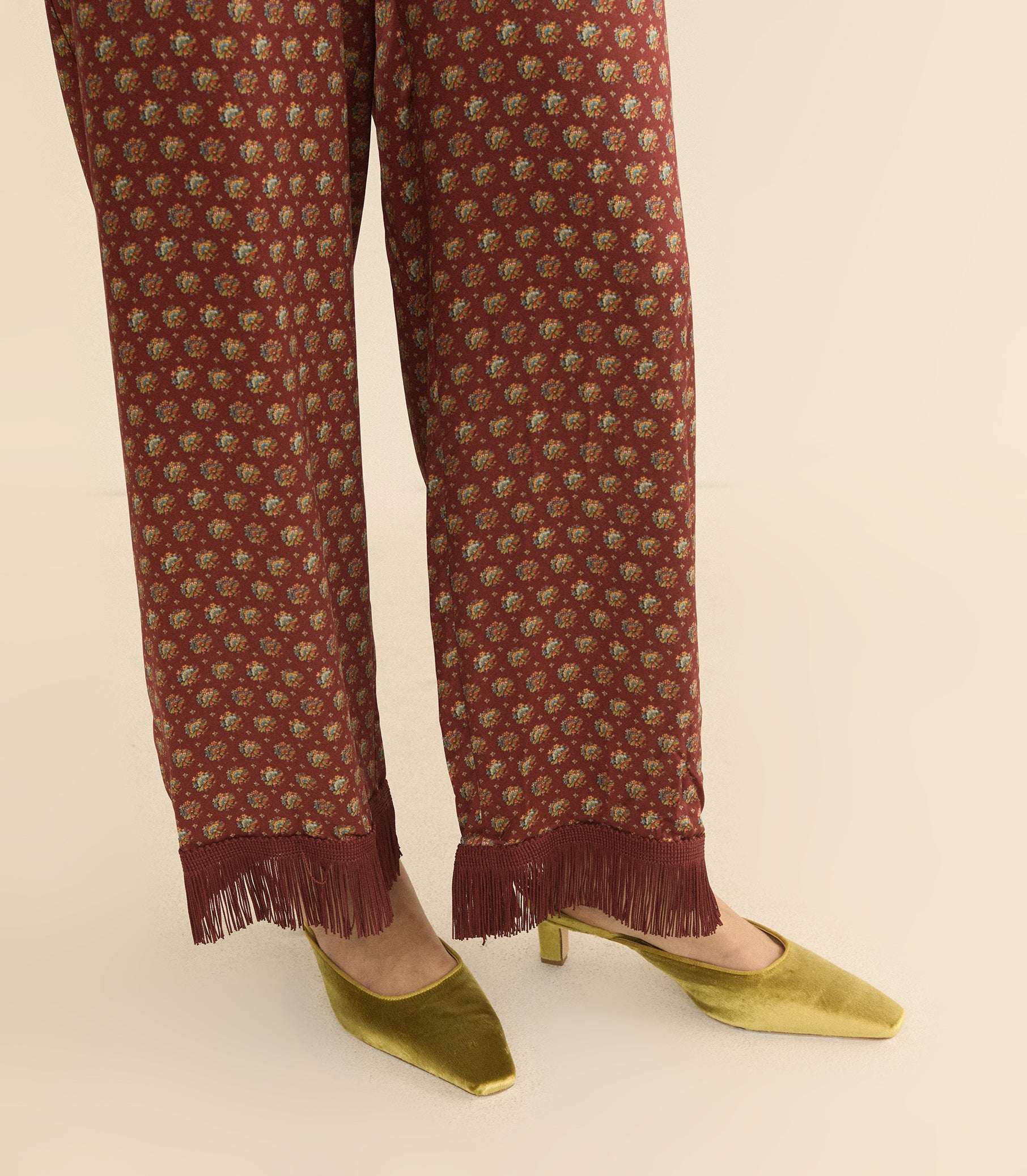 A close-up of a person's legs wearing rust-brown wide-leg trousers with a small floral pattern and fringe hems, paired with gold pointed-toe heels on a light beige background. 1
