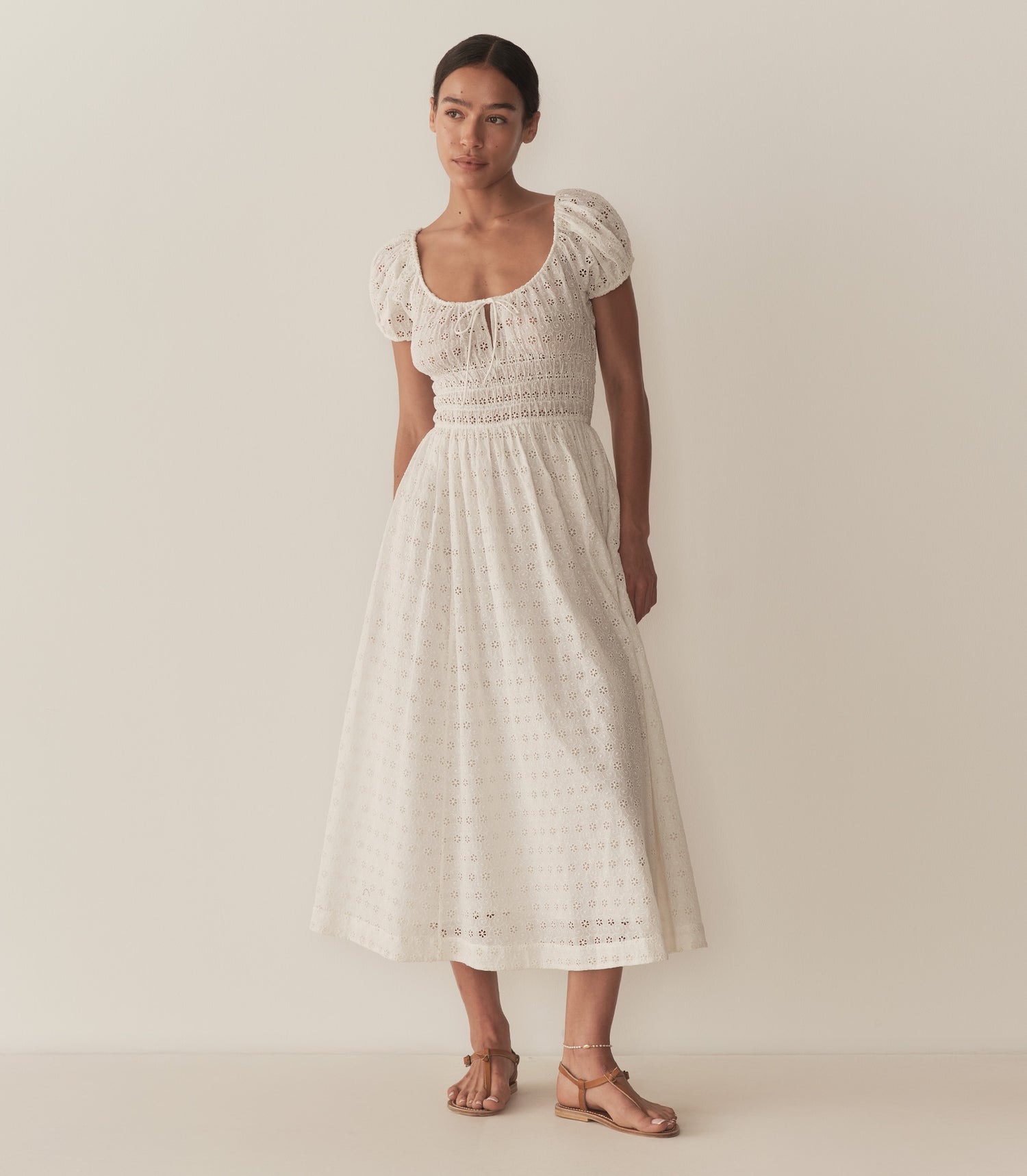 Zahara wears a long white eyelet dress with short puff sleeves, paired with tan sandals.