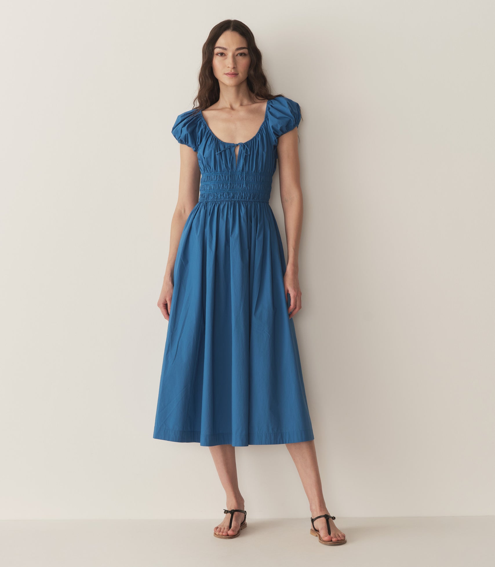 Bruna is wearing a long blue cotton dress with short puff sleeves and sandals.