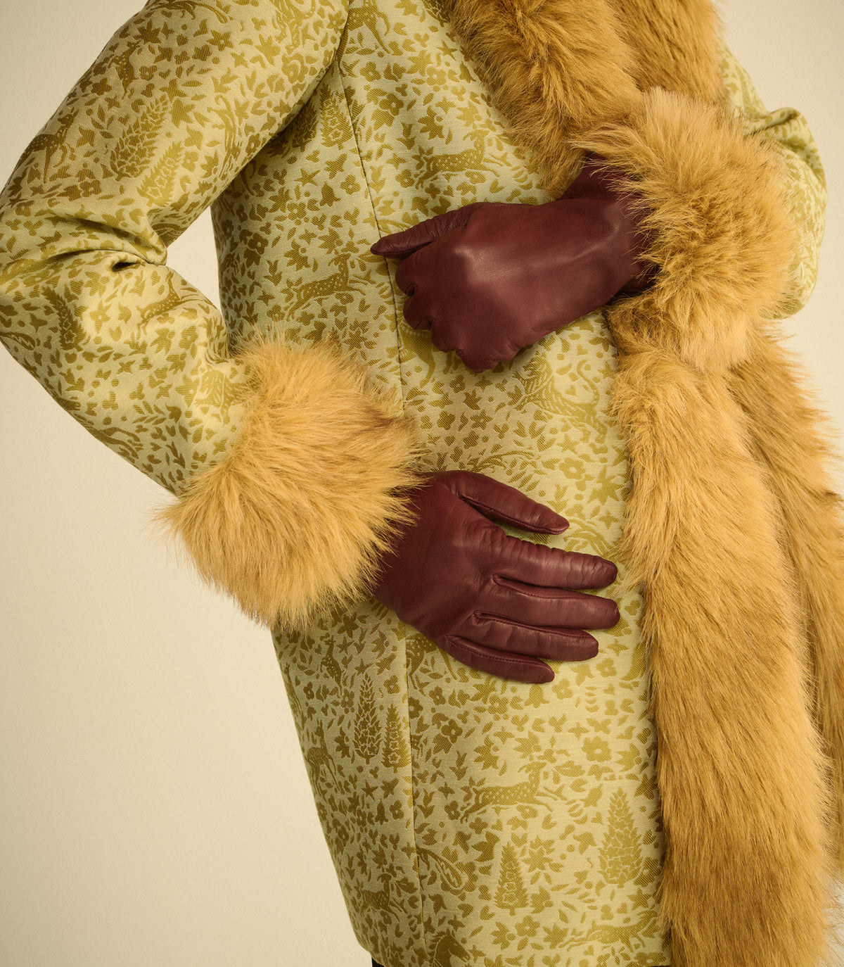 A close-up of a person wearing a long olive-green patterned coat with tan faux-fur trim and dark burgundy leather gloves, posed against a plain light beige background.