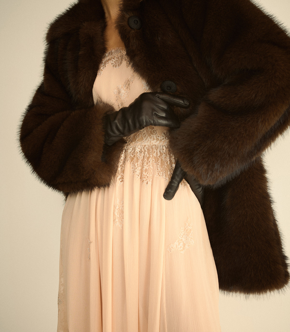 A close-up of a person wearing a dark brown fur jacket over a pale blush pink dress with lace detailing, black leather gloves, and a pearl necklace, set against a plain light beige background.