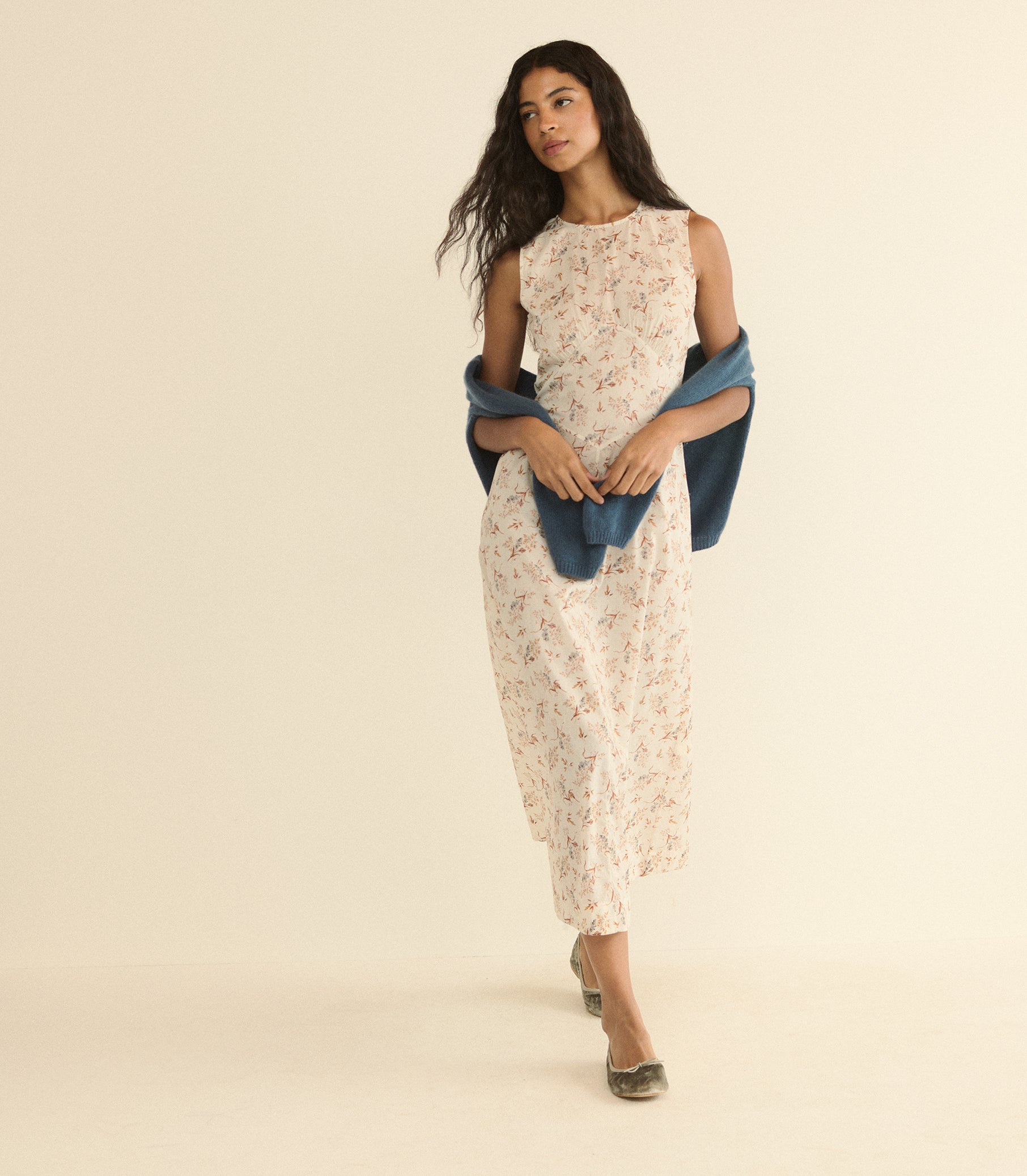 A woman modeling a sleeveless cream dress with a delicate floral print, styled with a draped blue shawl and metallic flat shoes, set against a solid warm beige studio background.