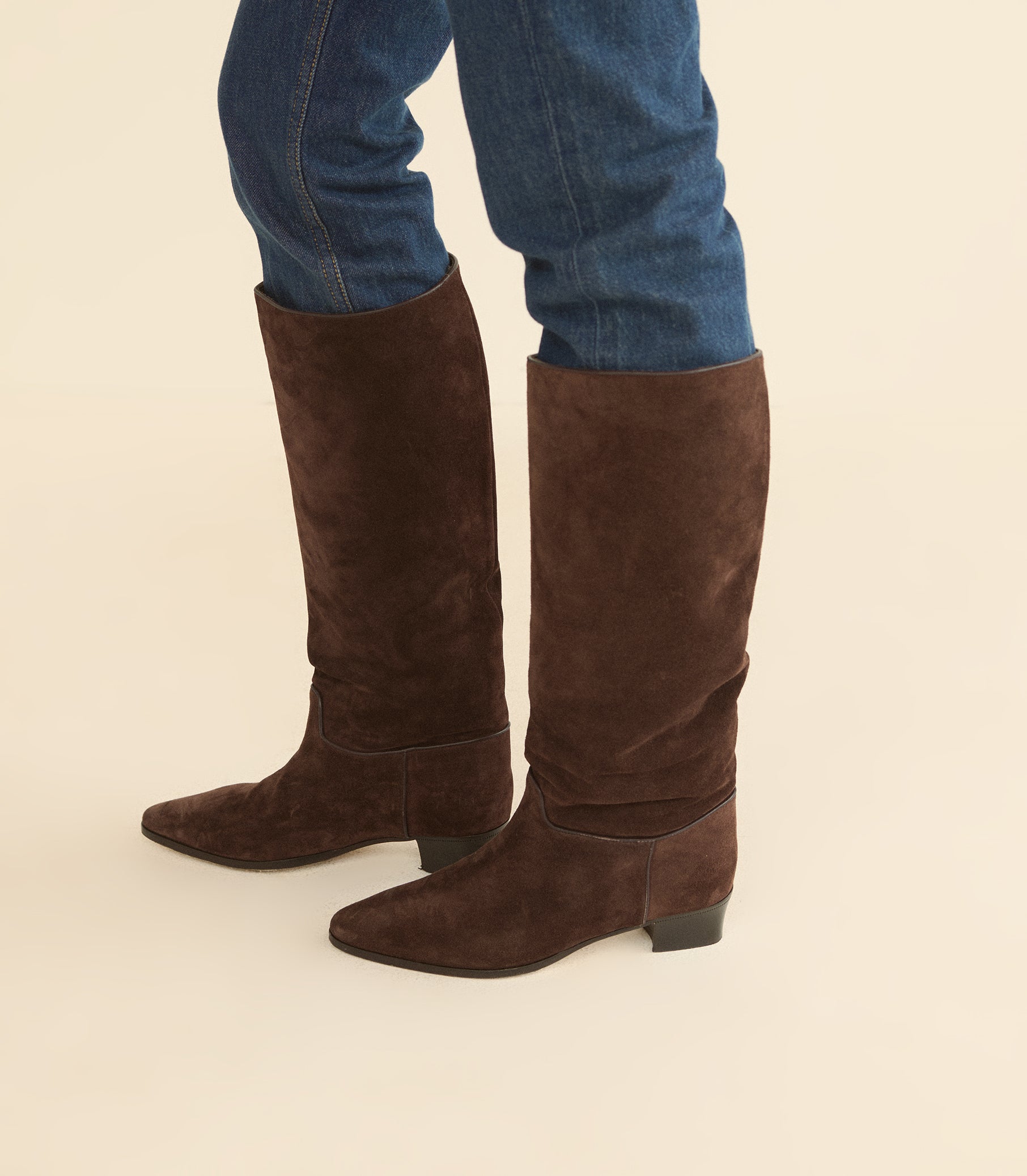 A pair of dark brown suede knee-high boots worn with blue jeans, shown from the knees down against a solid light beige background.