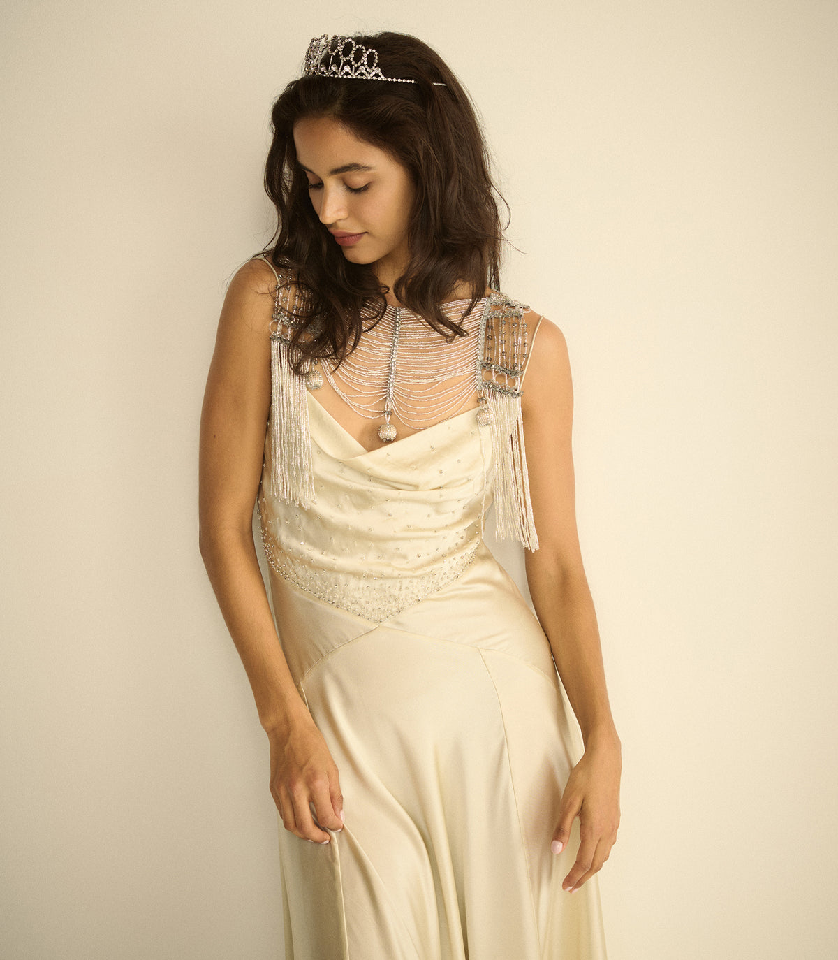 A woman wearing a cream-colored 1920s-style flapper gown with silver beaded shoulder straps, a matching beaded collar, and a silver tiara, posed against a plain beige background.