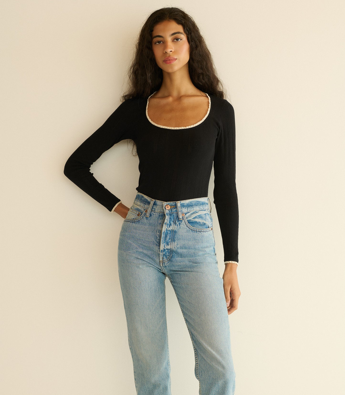 Woman wearing a black long-sleeve top and light blue jeans against a plain background